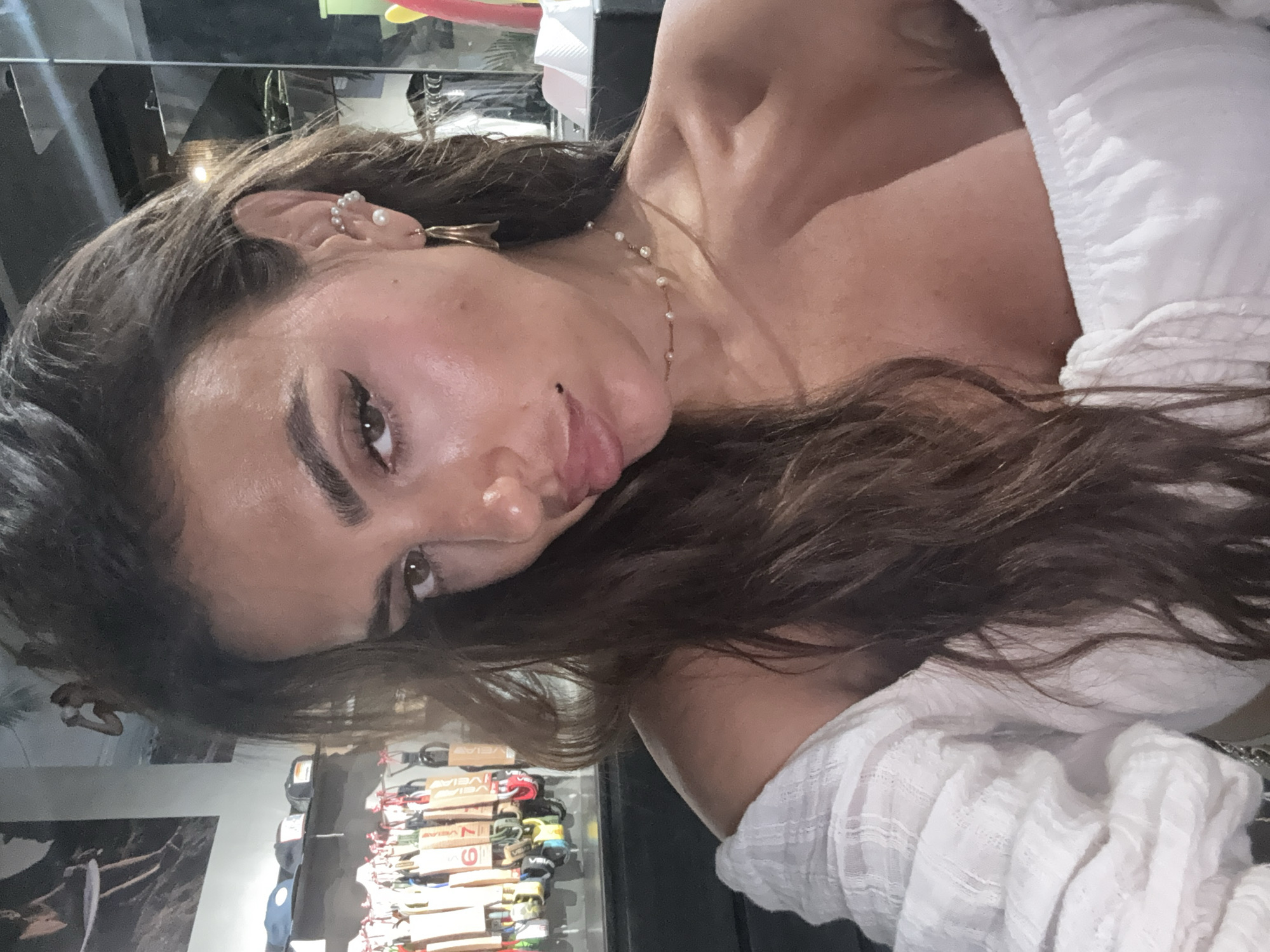 A woman with long brown hair is shown in an eye-level shot. She is wearing a white, textured top and a delicate necklace with small beads. She has a variety of earrings in her left ear. The background appears to be a retail environment with shelves of items, possibly shoes or related merchandise. The location is identified as North Kuta, Indonesia.