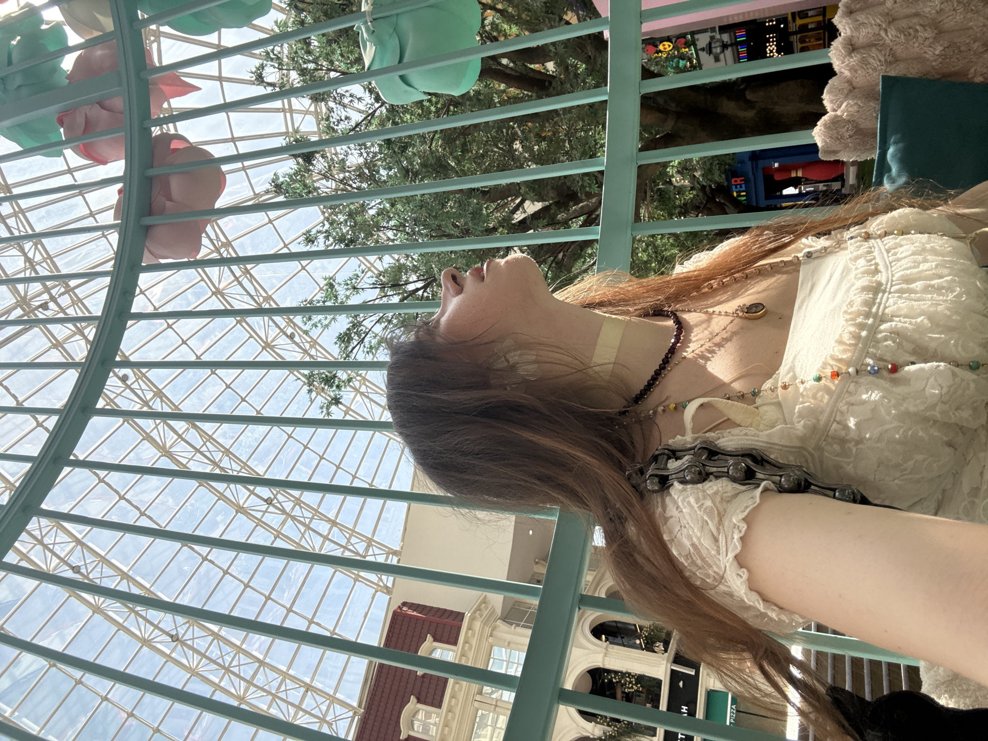 The image is taken from a low angle, looking up, seemingly through a green metal railing or structure. A person with long brown hair is positioned within the frame, leaning back against the structure. The person is wearing a white, lace-trimmed top and necklaces. Above, there is a large glass roof structure with a grid pattern. In the background, there are building facades with architectural details, a tree with green foliage, and hanging decorations. The location appears to be a brightly lit indoor space, possibly a shopping mall or atrium. While specific details about the city of Moscow are not readily apparent, the architecture in the background is consistent with the style found in Moscow, Russia.