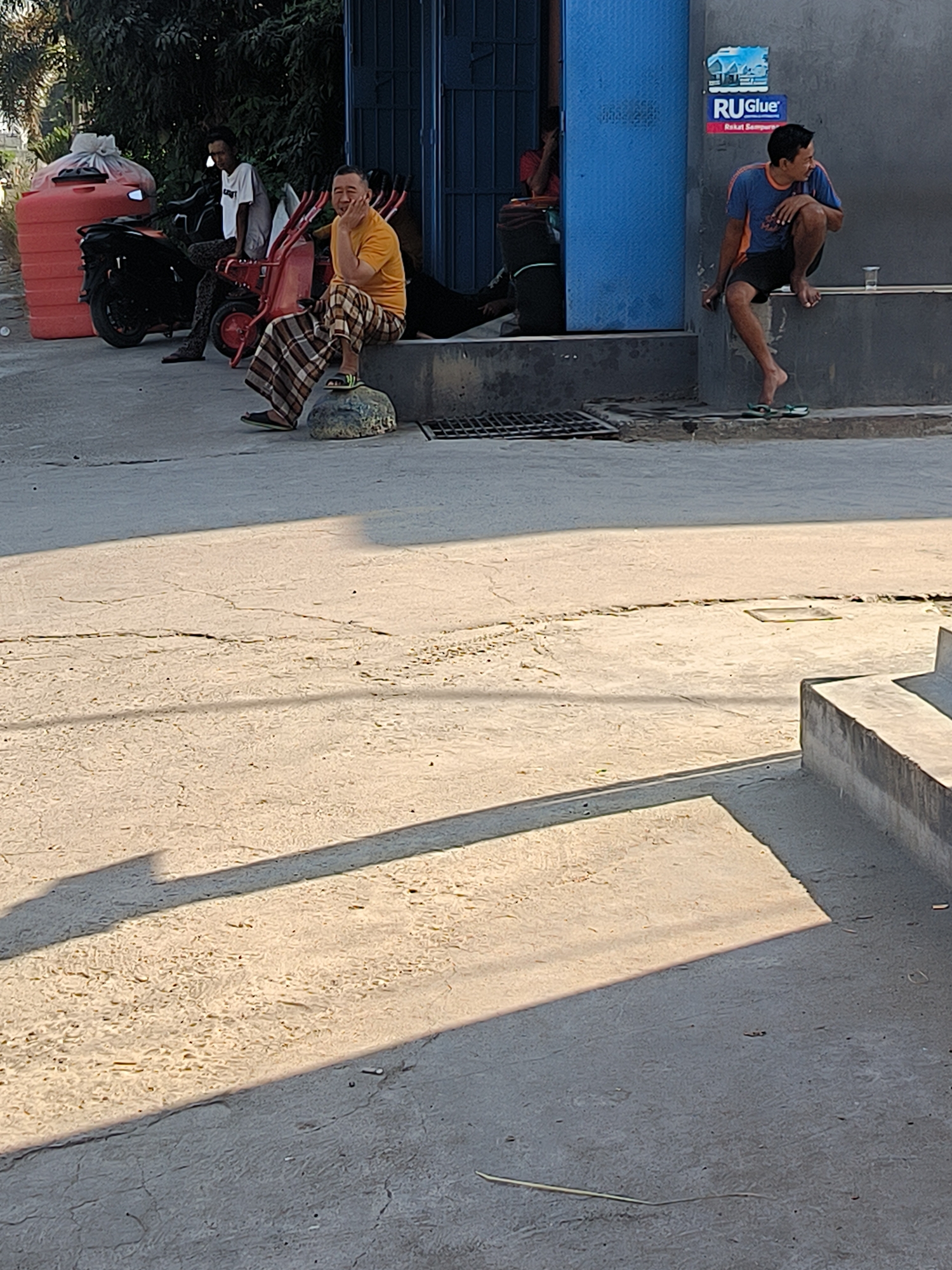 In Bumi Mulyo, Indonesia, two men are visible outdoors. One man is seated on a concrete ledge, wearing a blue and orange t-shirt and black shorts, with his legs dangling and a plastic cup on the ledge beside him. Another man sits on a rock, facing right, wearing a yellow t-shirt and plaid pants, with his hand resting on his chin. To the left, another man is seated on a red cart, wearing a white t-shirt and patterned pants, with a black motorcycle parked behind him and a large red water tank to the left. The environment consists of a concrete ground, a building with a blue door and a gray wall with a RU.GLUE advertisement, and trees in the background. The scene is illuminated by sunlight, casting long shadows on the ground.