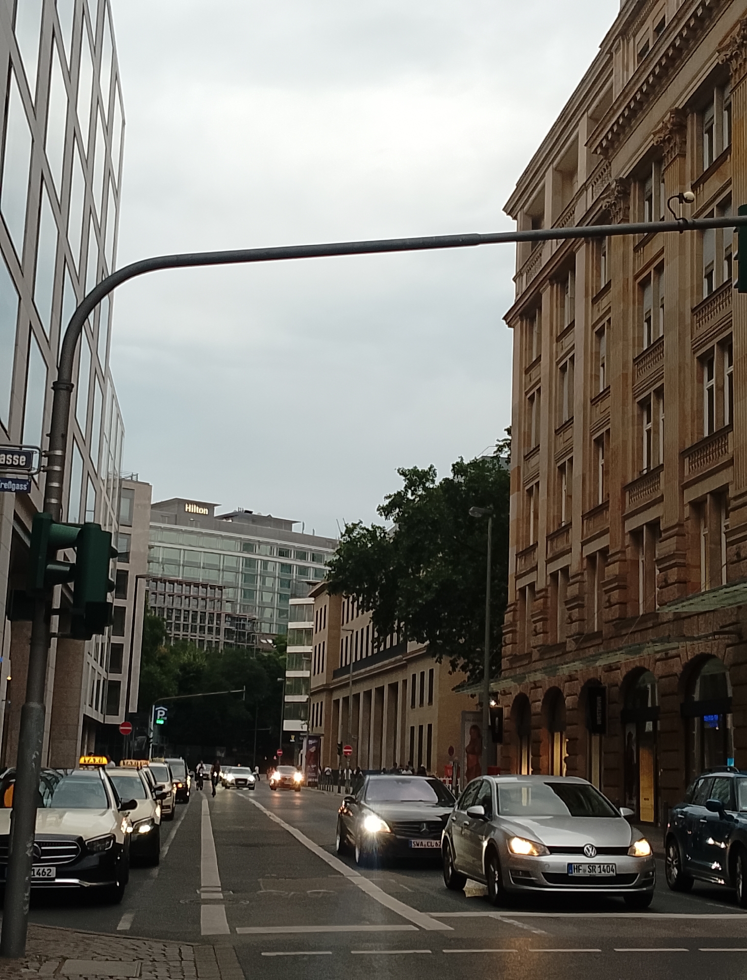 A medium shot captures a street scene with a clear sky, suggesting daylight. On the left, a modern building with a glass facade dominates the foreground, its geometric patterns reflecting the sky. A traffic light, showing green, is mounted on a pole in front of this building, along with a street sign reading "Gasse". To the right of this, a tall, tan building with classical architectural elements, featuring columns and arched windows, rises into the frame. A contemporary Hilton hotel with a glass facade is visible in the middle distance, between the two buildings. The street itself is paved with asphalt and marked with white lines. Several cars are on the road, including taxis with illuminated "TAXI" signs. One car in the foreground has license plate number "1462", and another has "HF SR 1404". Pedestrians can be seen on the sidewalk in the distance. Trees line parts of the street, and the overall atmosphere suggests an urban environment.