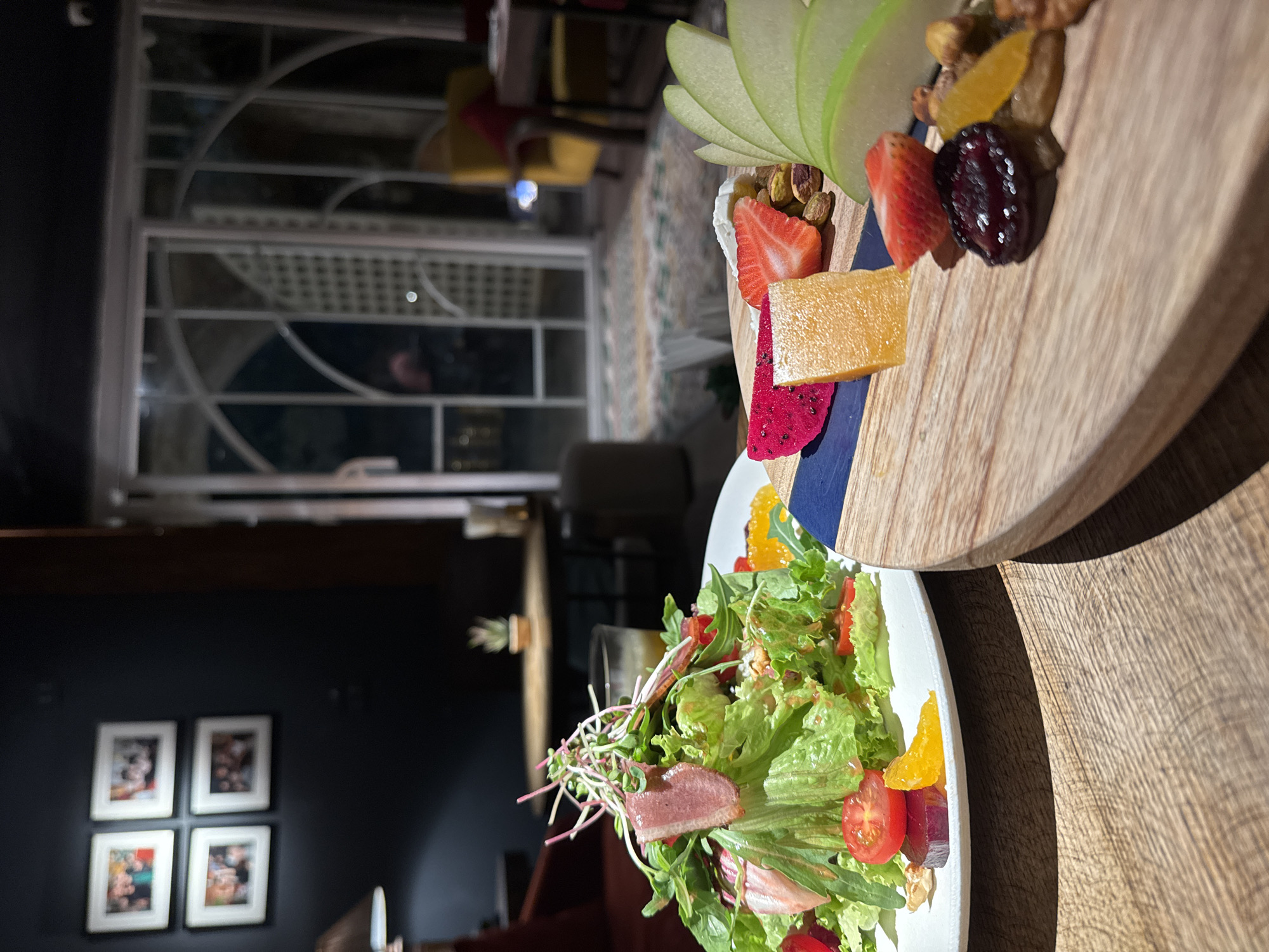 This image shows a close-up of two plates of food on a wooden table, likely in a restaurant setting in Hanoi, Vietnam. The foreground features a salad with lettuce, tomatoes, orange slices, and what appears to be slices of ham or cured meat. Microgreens are also visible, adding a decorative touch. In the background, a wooden platter displays a selection of fruits and nuts, including sliced green apples, strawberries, dried cranberries, pistachios, and dried apricots, along with a piece of cheese. The lighting suggests it could be evening or late afternoon. The overall scene is one of a meal being presented, with an emphasis on fresh ingredients and appealing presentation. The room appears to have dark walls adorned with framed pictures, and a glimpse of a window with intricate lattice work can be seen in the blurred background, possibly indicating a traditional architectural element. There is no visible text in the image.