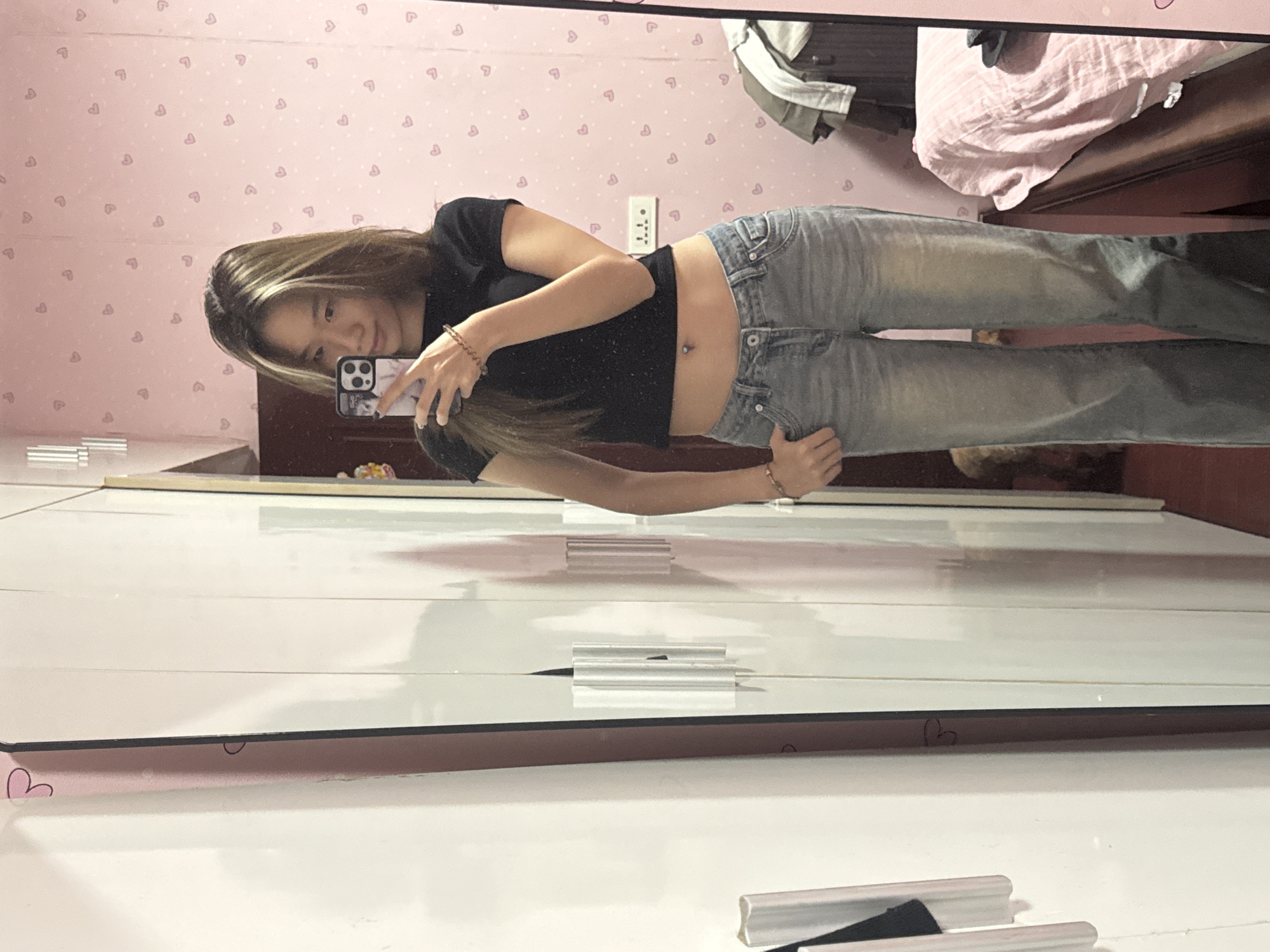 A person is taking a mirror selfie indoors. The individual is fair-skinned with long, light brown hair and is wearing a black cropped t-shirt and light blue jeans. The person is holding a smartphone in their right hand, angled towards their face. A belly button piercing is visible. The background shows a pink wall with a pattern of small hearts, an electrical outlet, and a bedroom with a bed covered in pink and white linens. The reflection in the mirror reveals part of a white dresser or wardrobe. The photo was taken indoors, likely in a bedroom. There is no verifiable information from the image to suggest that this photo was taken in the city of Shapingba District, China.