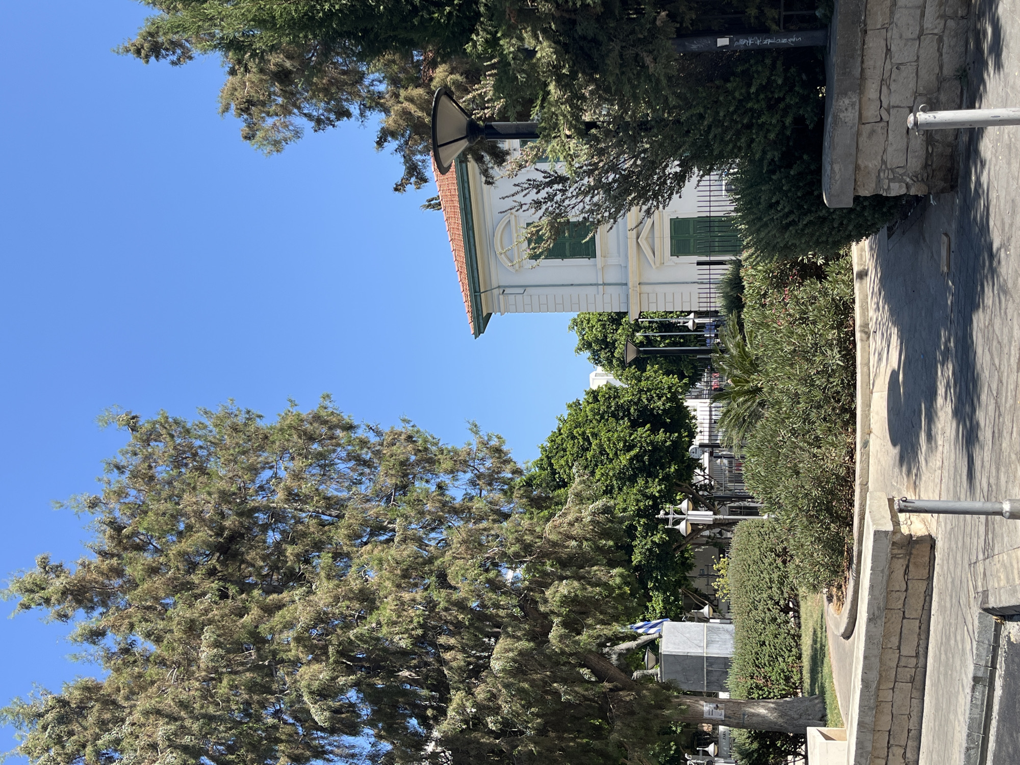 This is an eye-level, medium shot on a sunny day showing a view of a white neoclassical building with green shutters and a terracotta roof, surrounded by lush greenery and trees. The building, with its distinct architectural features, is partially obscured by trees in the foreground and to the right. In the foreground, there is a paved walkway and a low stone wall. A lamppost with a modern design stands to the right, silhouetted against the greenery. A statue with a Greek flag is visible to the left, near a large tree. The background is a clear blue sky. The scene is located on Gladstone Street in Limassol, Cyprus, in the vicinity of the Municipal Arts Centre. There are no people visible in the frame.