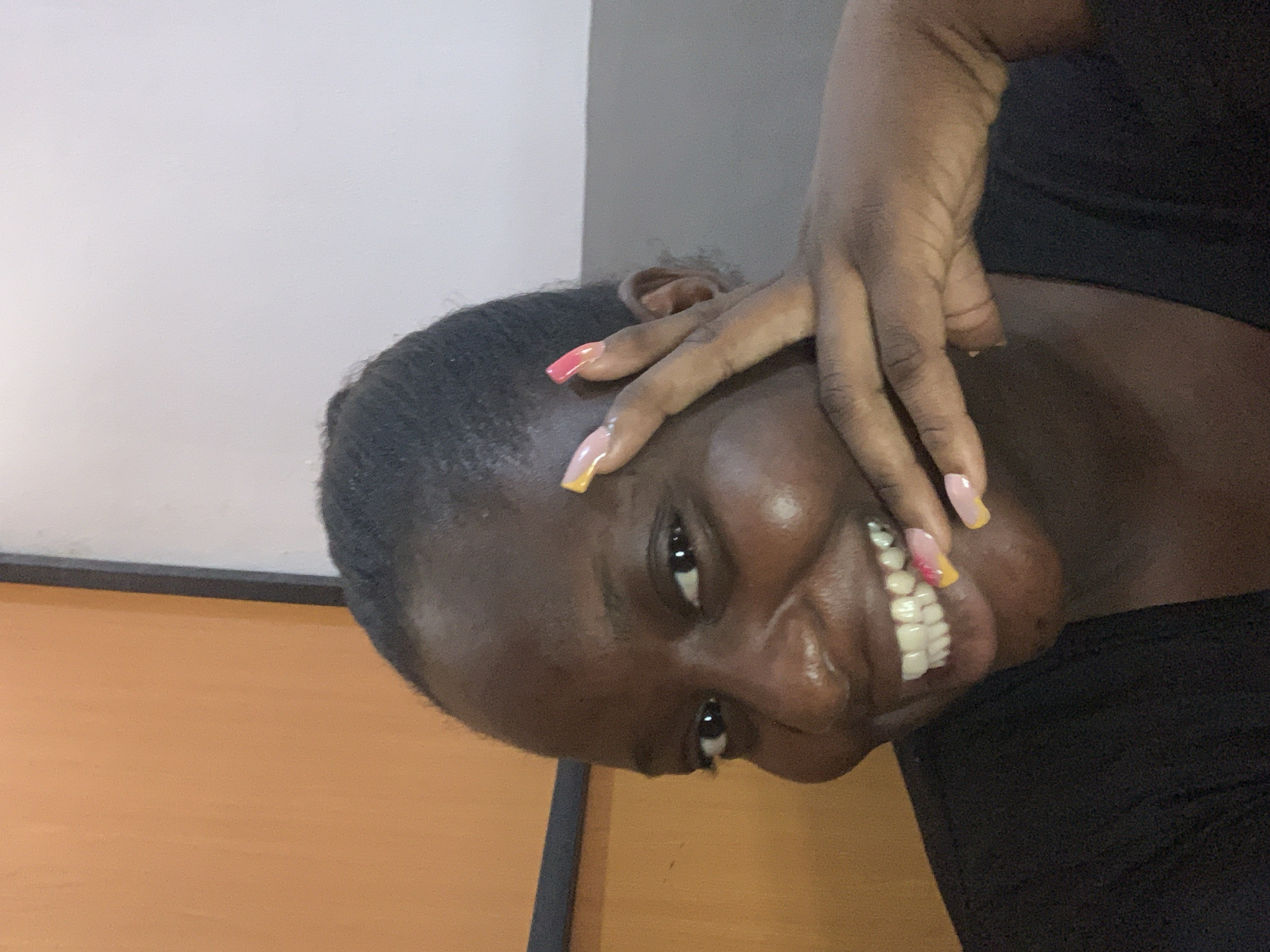 A close-up, eye-level medium shot shows a Black woman from the chest up, looking at the camera with a wide smile. She has short, dark hair and her hand is near her face, with her fingers extended. Her nails are long and painted with a gradient of pink and yellow. The background is divided into two sections: on the left, an orange wall, and on the right, a white wall. A dark frame runs vertically between the two walls, with a gray stripe below the white wall on the right.