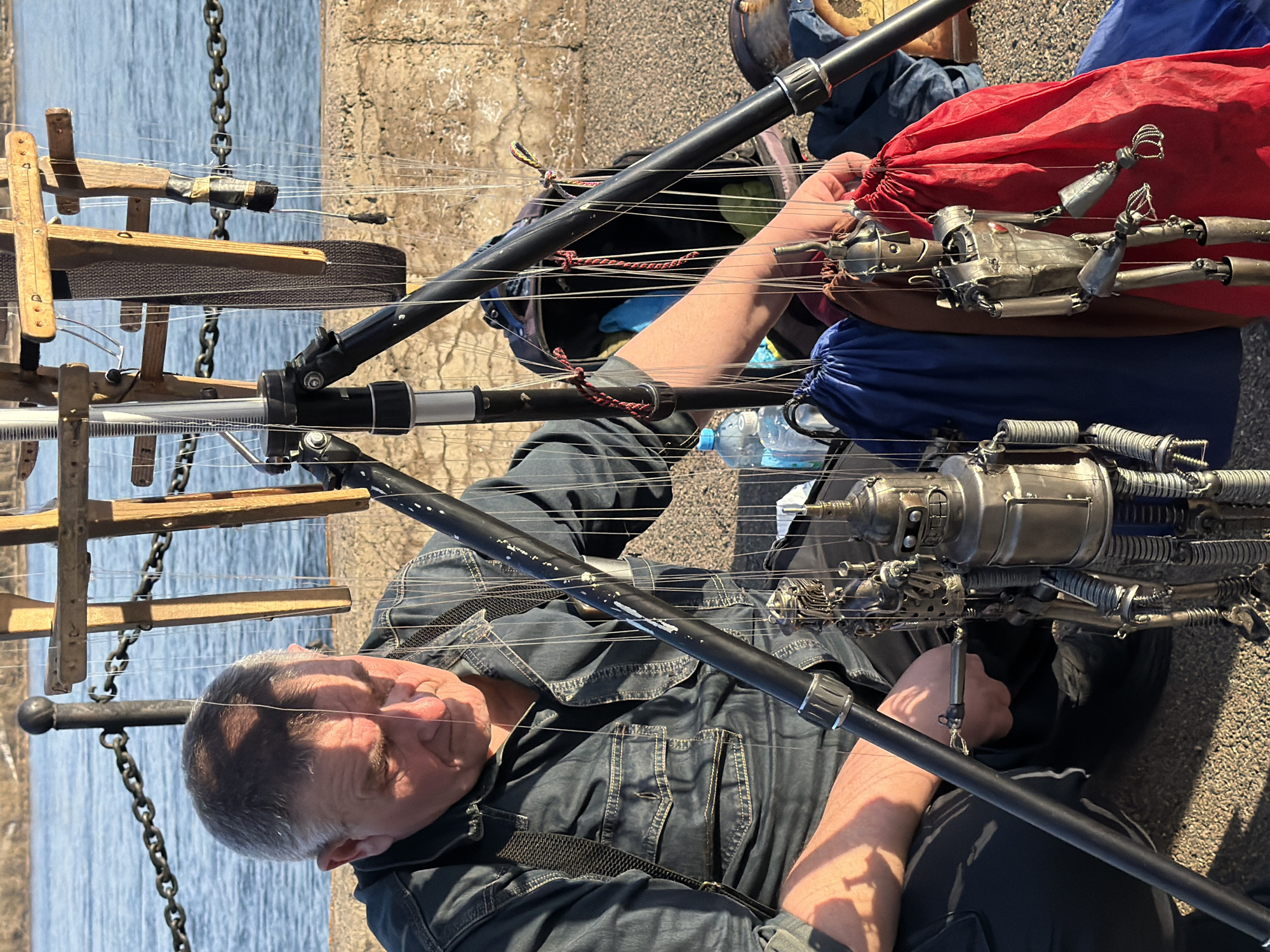 A man is sitting on a concrete surface with metal string puppets laid on the ground and a lake in the background. The man wears a denim shirt and has gray hair with a mustache. There are multiple metal string puppets, one partially inside a red drawstring bag. Puppet control bars sit to the left, with chains hanging near them. A water bottle and backpack are located next to the man. The concrete ground is visible in the foreground and background, and the location is Krasnoyarsk, Russia.