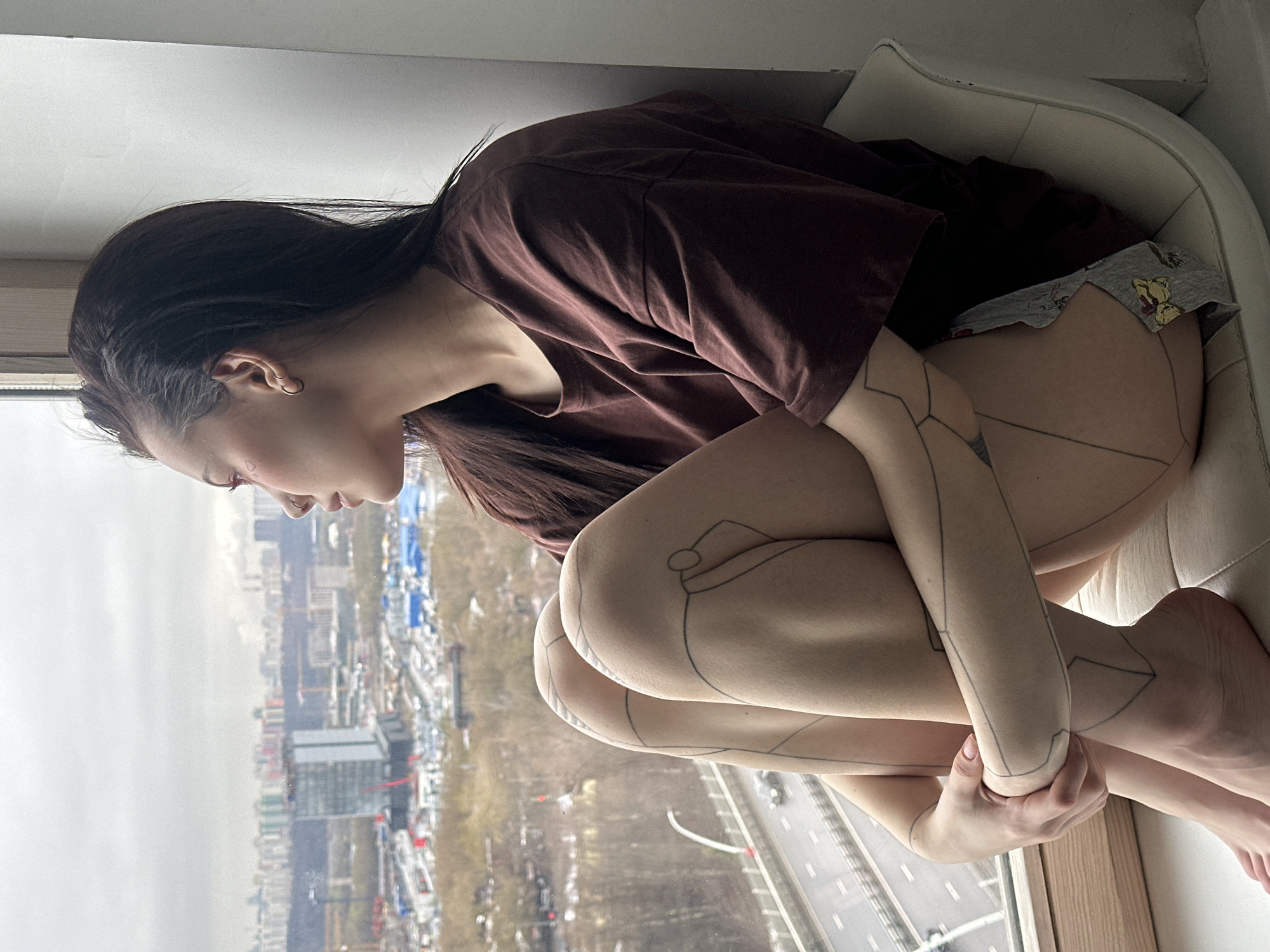 A young woman is sitting on a window seat in a building in Khimki, Russia. She is wearing a brown t-shirt and patterned underwear. Her knees are drawn up to her chest and her arms are wrapped around her legs. She has tattoos on her arms and legs. Outside the window is a cityscape with buildings, trees, and a highway. The sky is overcast.