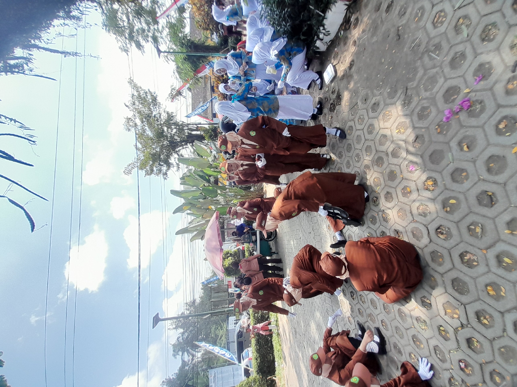 In Genteng Kulon, Indonesia, a group of individuals, likely students wearing matching brown uniforms and caps, are gathered outdoors on a paved area with hexagonal interlocking stones. Some are kneeling or crouching on the ground, while others are standing. A few individuals in the background wear blue and white attire and headscarves. The environment appears to be a public space with trees, foliage, a pink umbrella, parked vehicles, and utility poles with overhead wires. The sky is visible above, with clouds and sunlight indicating it is daytime.