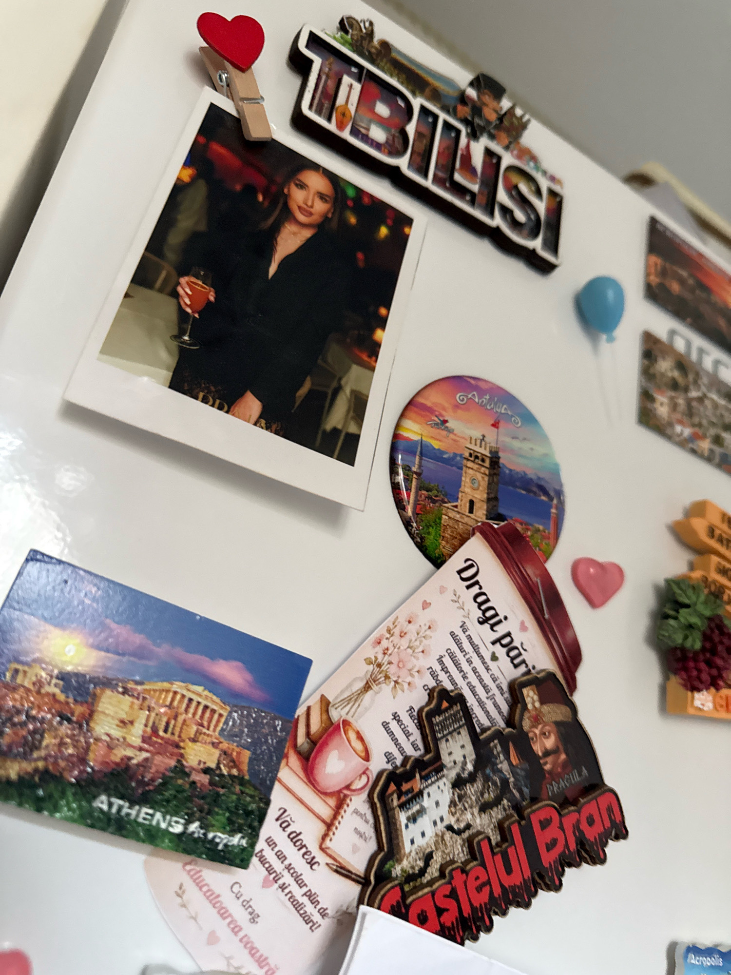 The image is a close-up, high-angle shot of a white refrigerator door adorned with various magnets and a photograph. The primary focus is on a collection of travel-themed souvenirs. To the upper left, a wooden clothespin shaped like a red heart holds up a printed photograph of a woman in a black dress holding a champagne flute. To the right of the photo is a wooden magnet with the word "TBILISI" in bold lettering, featuring smaller images of landmarks within the letters. Below the "TBILISI" magnet is a round, glossy magnet depicting a city skyline with a prominent clock tower and a body of water, identified by the text as "Antalya".
Below the Antalya magnet, there are several other magnets. One is a rectangular magnet with an image of the Acropolis in Athens, Greece, with the text "ATHENS" visible. To the right of the Athens magnet is a magnet featuring a castle and a portrait of Dracula, with the text "Castelul Bran" written in a dripping, blood-like font. Another magnet, partially visible, appears to be a bunch of grapes. There is also a small, pink heart-shaped magnet.
A small blue balloon magnet is positioned near the top right of the image. Several other magnets, mostly obscured or out of focus, are visible along the top and right edges of the frame. The overall environment is a domestic setting, likely a kitchen, given the refrigerator. There are no people or actions depicted beyond the static display of souvenirs. The image is well-lit, with a slight glare on the refrigerator surface. The location is not specified to be in the city of Chișinău, Moldova; the magnets themselves represent various travel destinations.