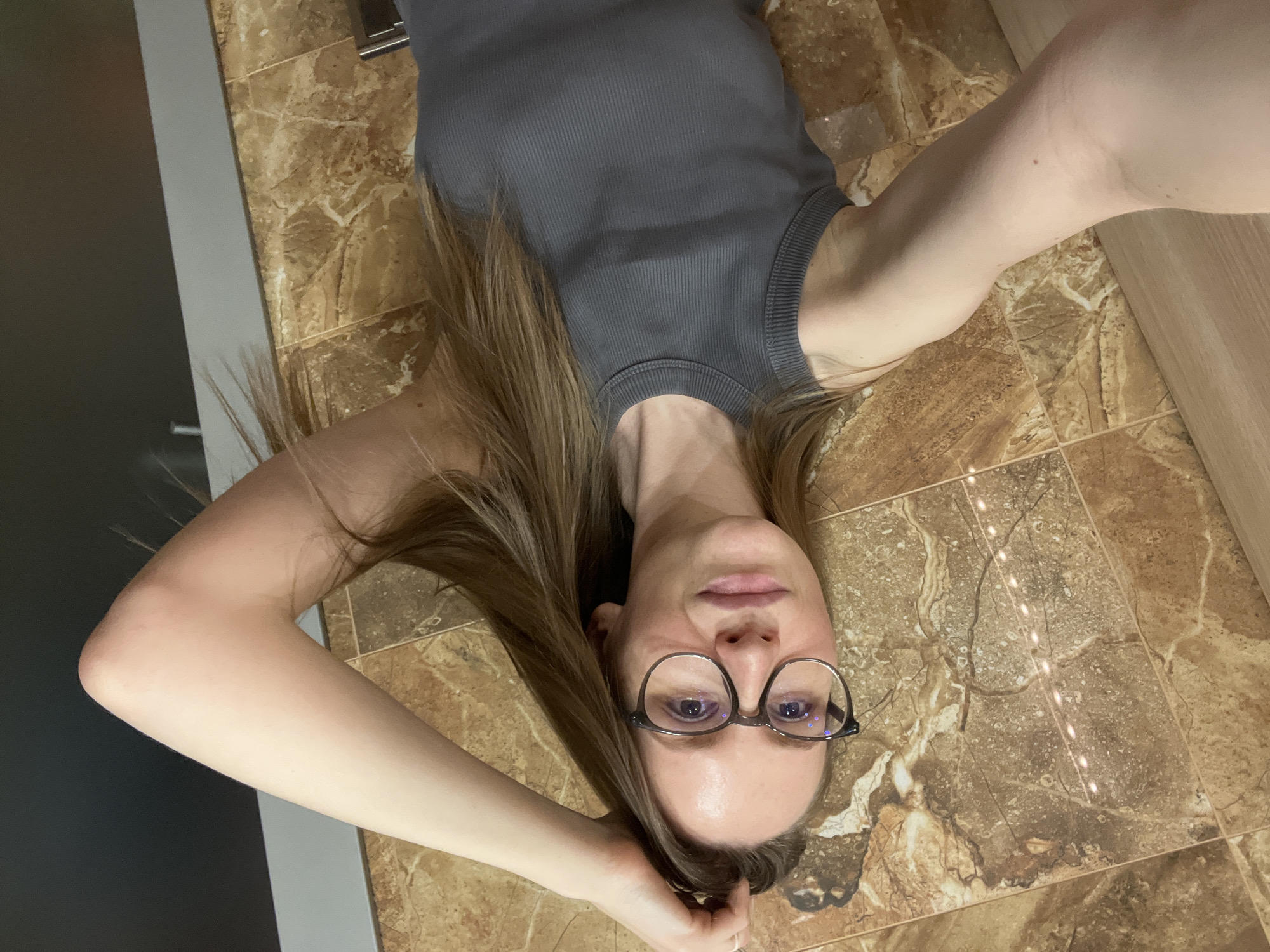 A young woman with long brown hair and wearing glasses and a gray tank top lies on a brown-tiled floor. Her arms are raised and positioned to take a selfie. The location is likely indoors, with a gray panel on the left and a wooden panel on the right. The location in Khimki, Russia is not discernible from the image.