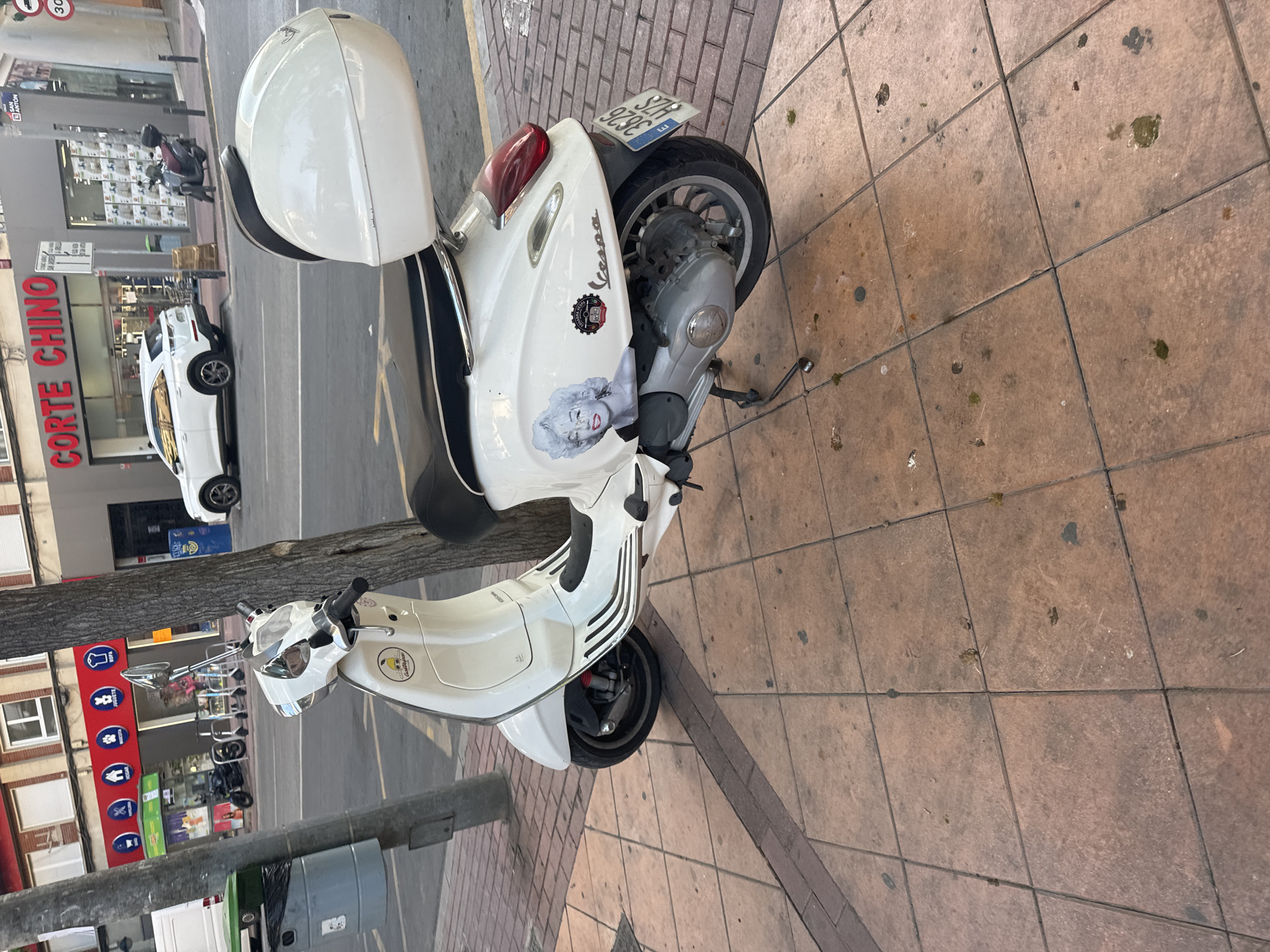 A white Vespa scooter is parked on a sidewalk paved with reddish-brown square tiles. The scooter has a white top box, a black seat, and a sticker of Marilyn Monroe on its side. A license plate with the numbers "3626 HZS" is visible on the rear. The scooter is leaning on its kickstand. Behind the scooter, a tree trunk with visible bark is present. In the background, a street can be seen with a white SUV parked and a store with the sign "CORTE CHINO" in large red letters. The store appears to be a retail establishment. The general environment suggests an urban setting. The image captures a street scene, likely in a city, with commercial activity and parked vehicles.