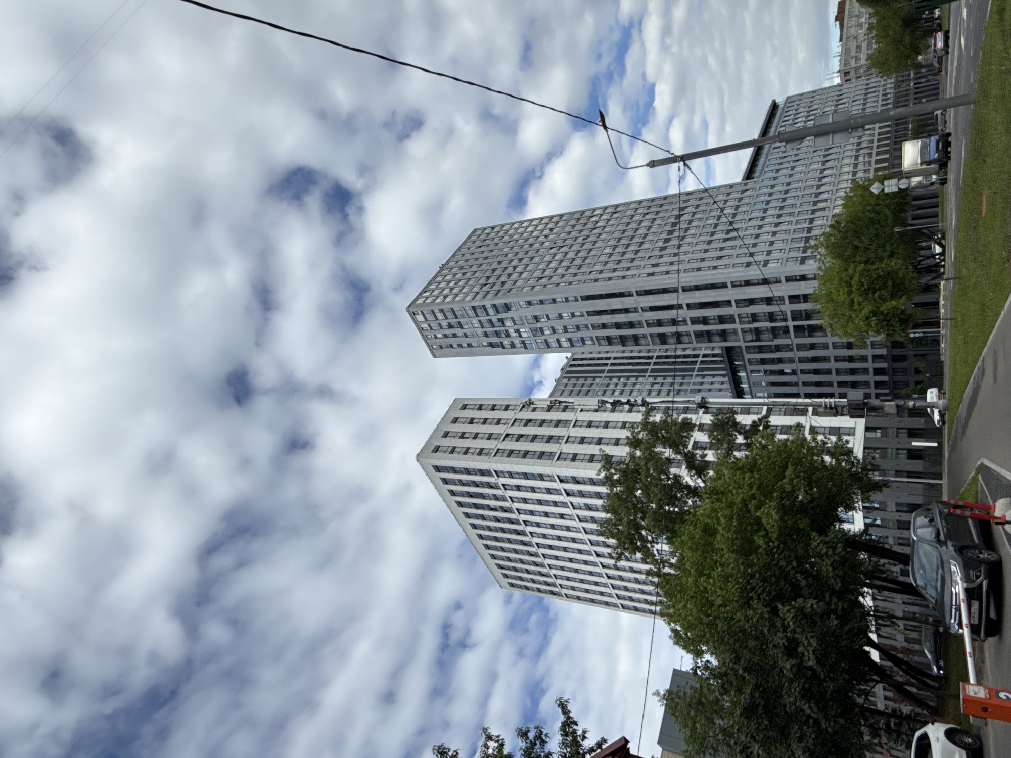 The image shows two high-rise buildings in Moscow, Russia, under a cloudy sky. The buildings are predominantly white with dark window patterns, suggesting a modern architectural design. Trees and greenery partially obscure the lower sections of the buildings, particularly on the right side of the frame. A street with parked cars is visible on the right edge of the image. Power lines cut across the sky in the upper portion of the frame.