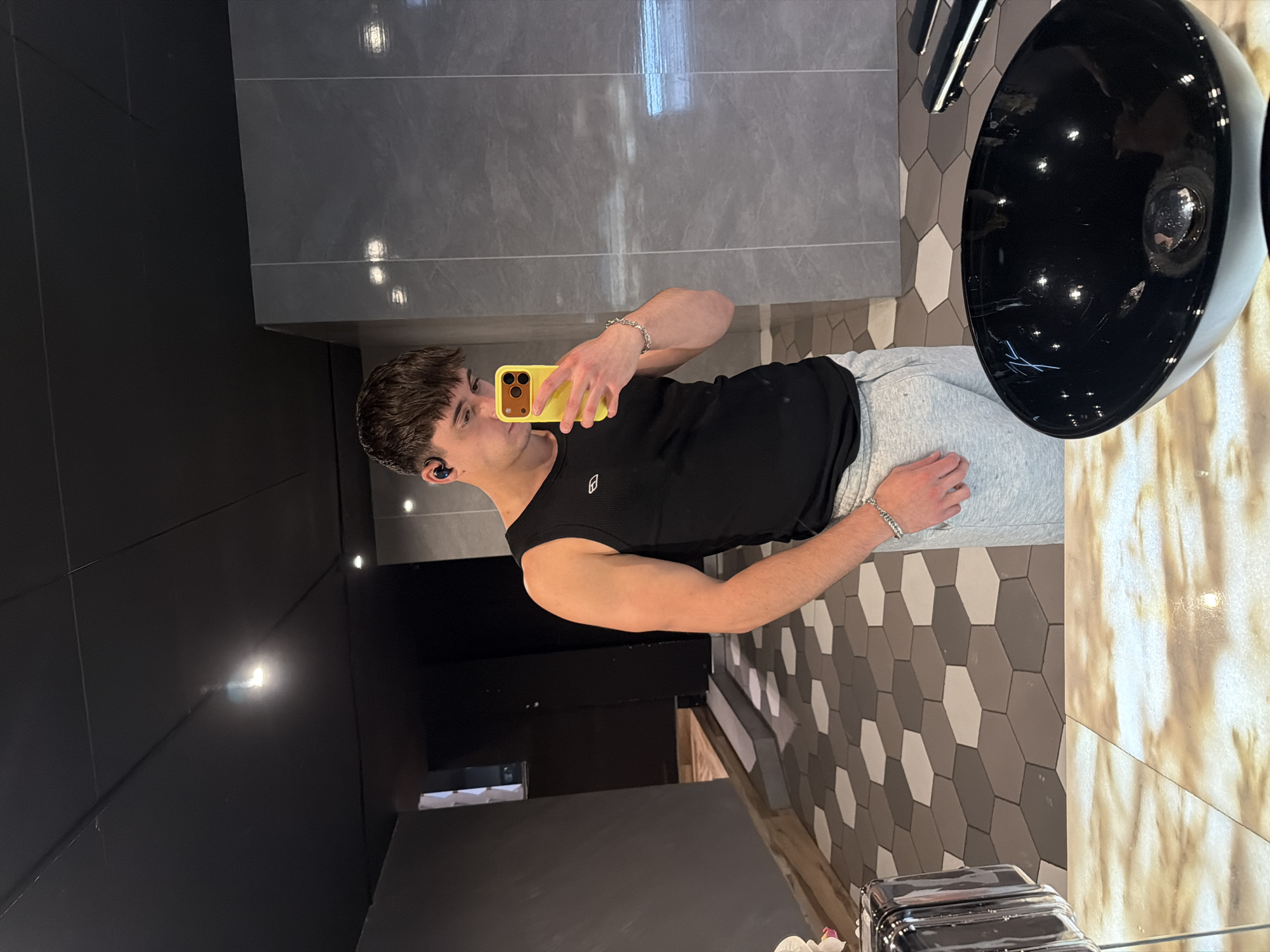 A young man is taking a selfie in a bathroom with a yellow phone. He is wearing a black tank top and gray sweatpants. The bathroom has a modern design with gray and white hexagonal tiles on the wall, gray tiles, and a black sink. The location is Sofia, Bulgaria.