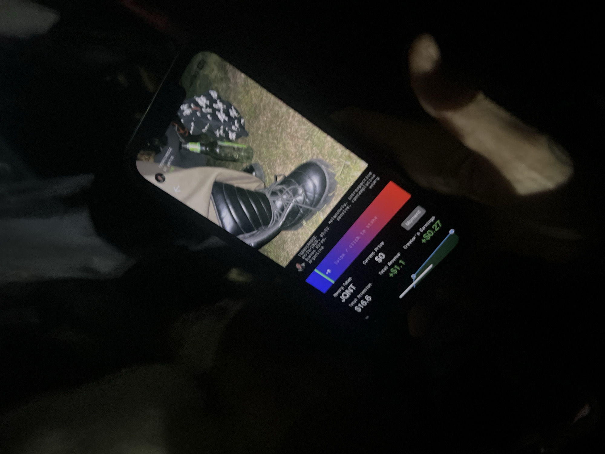 A hand holds a mobile phone displaying a video of an outdoor scene in Buenos Aires, Argentina, as identified by the on-screen text "TOPRUTAMOSE 08/28/2023, BUENOS AIRES ARGENTINA." The video shows a person in khaki-colored pants and black boots sitting on grass next to a black and white patterned garment and a green bottle. Below the video, the phone screen displays the words "melancholy, irrespective, pensive, contemplative, weary" and "swipe / click to stake." Also visible are "Current Price $0," "Total Revenue +$1.1," "Creator's Earnings +$0.27" and "Jonit Total Promotion $16.6."