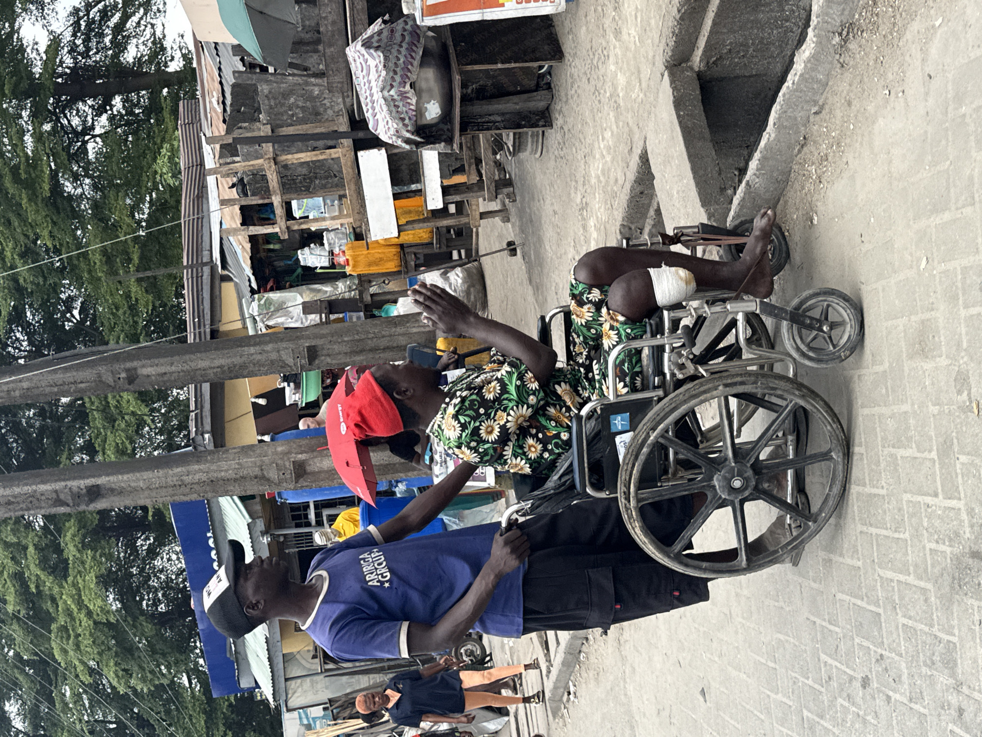 In a street scene in Ado, Nigeria, a man wearing a blue t-shirt and a black baseball cap pushes a wheelchair. Inside the wheelchair, a person wearing a red cap and a floral-patterned dress is seated, with a bandage around their left knee. The person in the wheelchair is clapping their hands. To the left of the wheelchair, another person wearing a dark blue polo shirt and light orange shorts walks away from the camera. In the background, market stalls and buildings are visible, with trees overhead. The ground is paved with grey bricks and concrete.