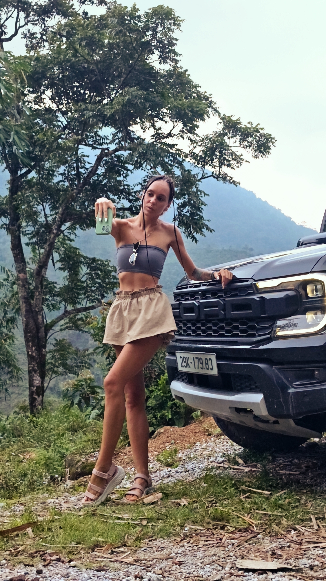 A young woman is captured in a full-body shot taking a selfie with a green smartphone, standing beside the front of a black Ford truck. She wears a grey bandeau top, light brown ruffled shorts, and light pink chunky sandals. Her dark hair is styled with small braids or twists, and sunglasses hang from her top. Tattoos are visible on her right arm.
The black truck, possibly a Ford Ranger or F-150 Raptor, features a large grille with "FORD" prominently displayed and modern headlights. A license plate, "29K-179.83," is affixed to the front bumper.
The scene unfolds outdoors in a natural, mountainous environment, characteristic of Nậm Dịch Commune, Vietnam. Tall green trees and dense foliage form the background, with hazy mountains visible in the distance under an overcast sky. The ground beneath her feet is unpaved, consisting of gravel and dirt with sparse grass. The lighting suggests it is daytime, possibly late afternoon, with soft, diffused light.