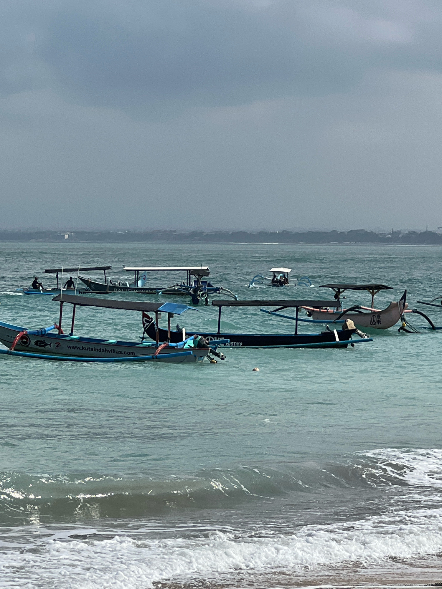 The image depicts a body of water, likely the ocean, with several traditional Indonesian outrigger boats anchored or moving on the surface. The water is a light turquoise color, and small waves are breaking near the shore in the foreground. The sky is overcast and gray.
In the foreground, there is a boat with a blue canopy and the text "www.kutaindahvillas.com" visible on its side, along with a fish logo. Another boat behind it, with a dark canopy, has the word "BROTHER" printed on its side. To the right of these, a light-colored boat with a pinkish hue features a peace sign and the words "ONE LOVE" painted on its stern. Further in the distance, other boats can be seen, some with people in them. One boat in the mid-ground appears to be labeled "BALI UNBEATABLE." The boats are characteristic of those used for transport and possibly tourism in coastal areas of Bali, such as Kuta.
The scene is consistent with the waters off the coast of Kuta, Indonesia, known for its beaches and boat activities. The presence of the website "kutaindahvillas.com" on one of the boats further suggests a connection to Kuta, as it is a popular tourist destination there. The overall impression is one of a calm, overcast day at sea, with multiple vessels engaged in various activities.