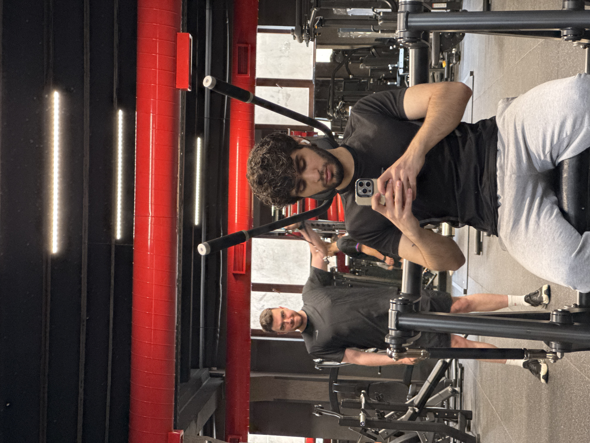 The image depicts a man sitting in a gym in Yerevan, Armenia, looking at his smartphone. He has curly brown hair, a beard, and is wearing a black t-shirt and light grey sweatpants. He sits on a seated lateral pulldown machine, looking at his iPhone in his hands. Behind him, another man wearing a black shirt and athletic shorts stands near the machine. The gym has red accents on the black walls.