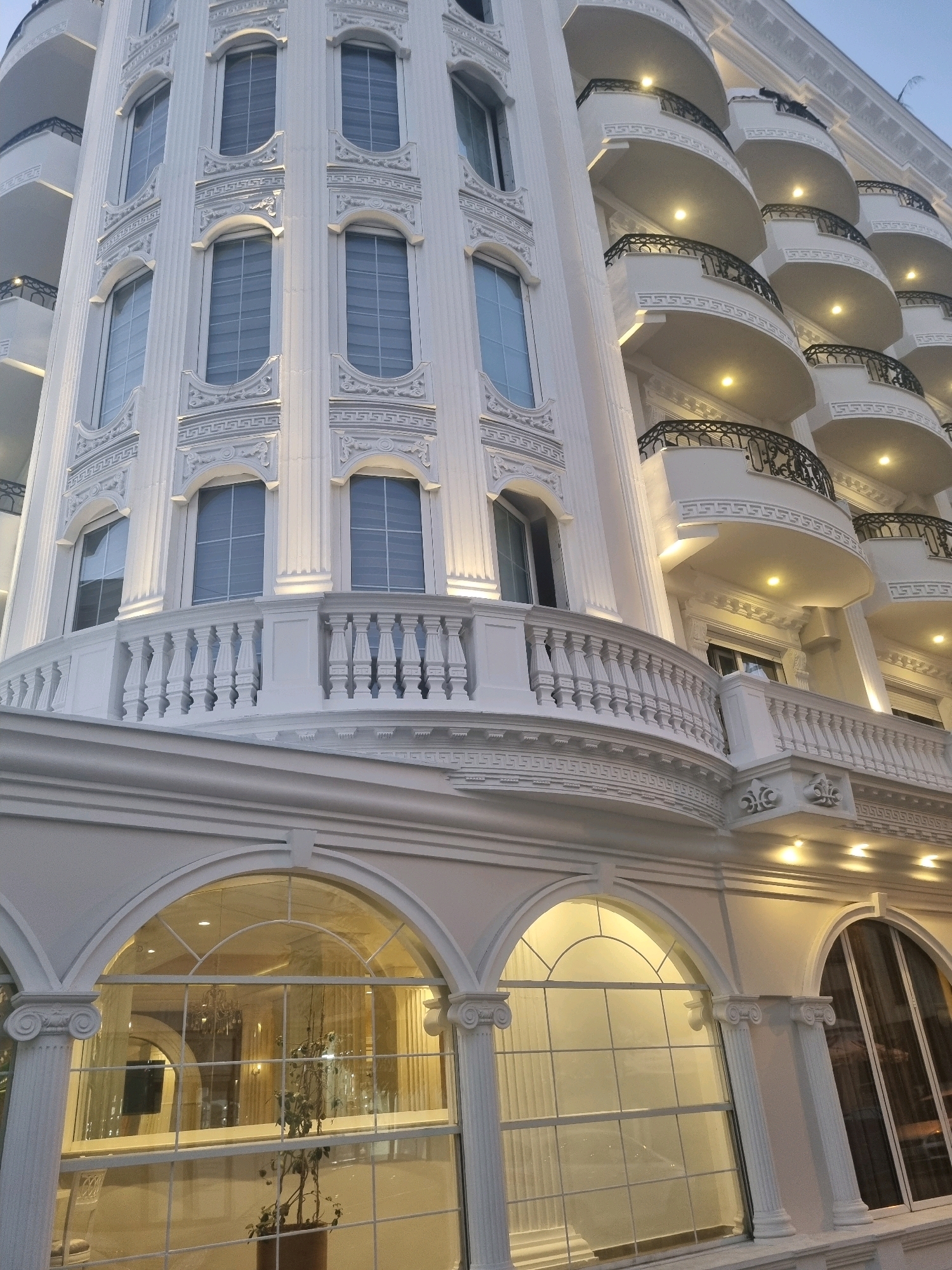 This is a low-angle, medium shot of a white building with ornate architectural details, likely a hotel or apartment complex, in Bashkia Durrës, Albania. The building features multiple levels of curved balconies with black wrought-iron railings, each illuminated by a spotlight. The facade is decorated with vertical fluting, decorative moldings, and arched windows with large panes. Below, arched glass entrances with classical columns are visible. The overall impression is one of grandeur and classical design. No people are visible in the frame. The sky is a pale blue, suggesting it is either early morning or late evening.