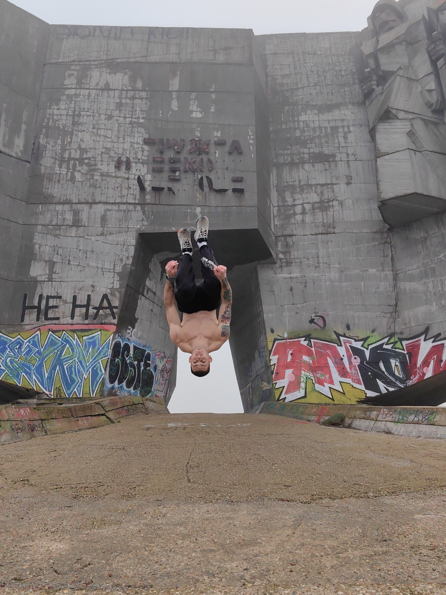 The image shows a man in the middle of a backflip in front of the Buzludzha Monument in Bulgaria, which is located in the city of Varna. He is upside down with his head facing down towards a concrete ground. He is wearing black pants and socks and shoes. He is shirtless and has tattoos on his arms. The monument is large and grey and covered in graffiti, especially on the sides where the concrete meets the ground. The weather appears to be foggy.