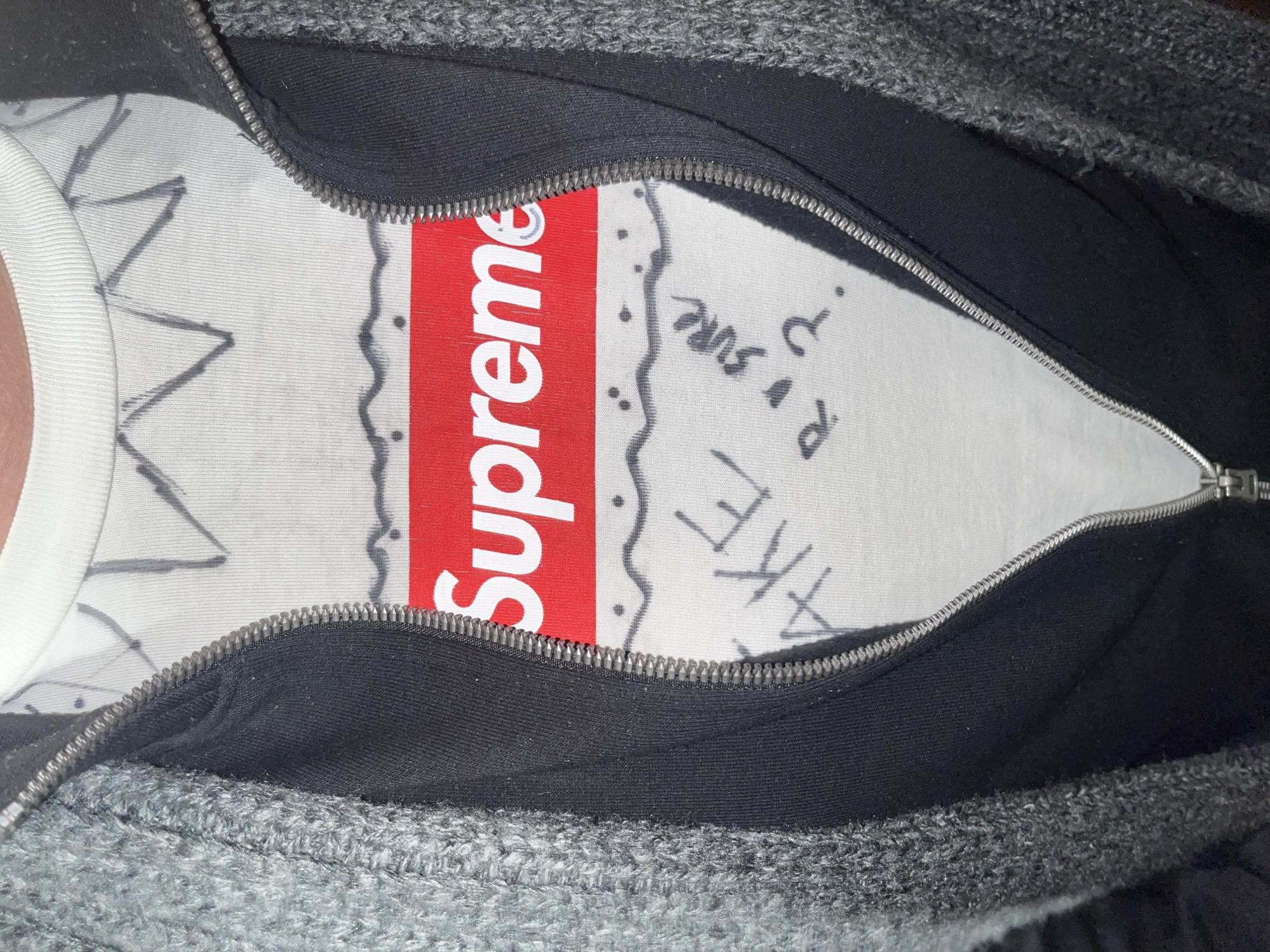 A close-up, medium shot depicts a person wearing a white T-shirt with a red "Supreme" logo partially visible. The shirt also features black line drawings and writing. The person is wearing a zippered sweatshirt in shades of gray and black. The sweatshirt is unzipped, revealing the shirt. The setting is unspecified, and there is no information to determine if the image was taken in Milan, Italy.