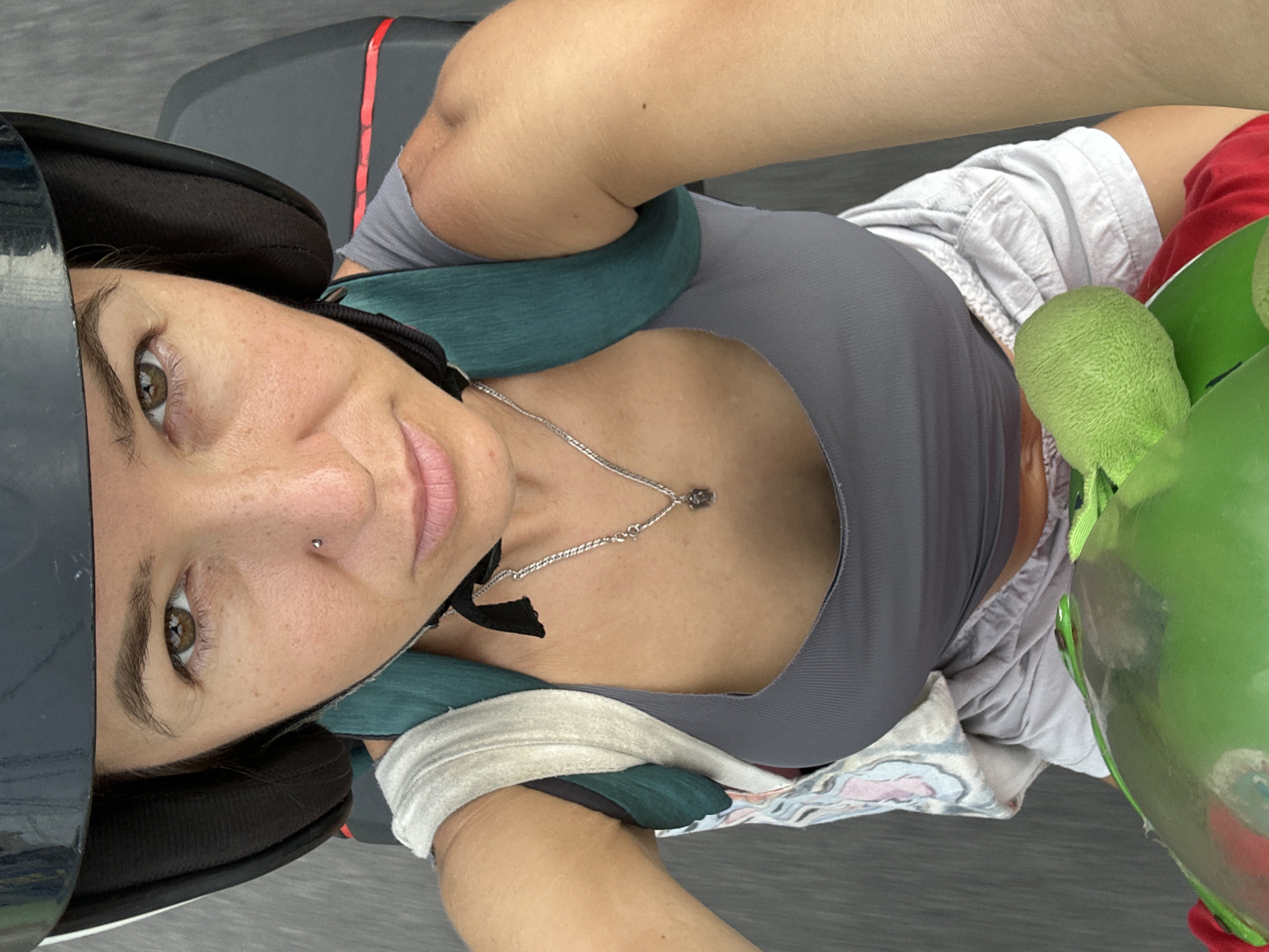 A woman wearing a black helmet and a grey tank top, with a backpack on, is riding a scooter on a paved road in Bac Nha Trang Ward, Vietnam. She has a nose ring and is wearing a silver necklace with a small pendant. The road appears to be grey asphalt. The image is taken from a first-person perspective, looking down at the woman and her surroundings.