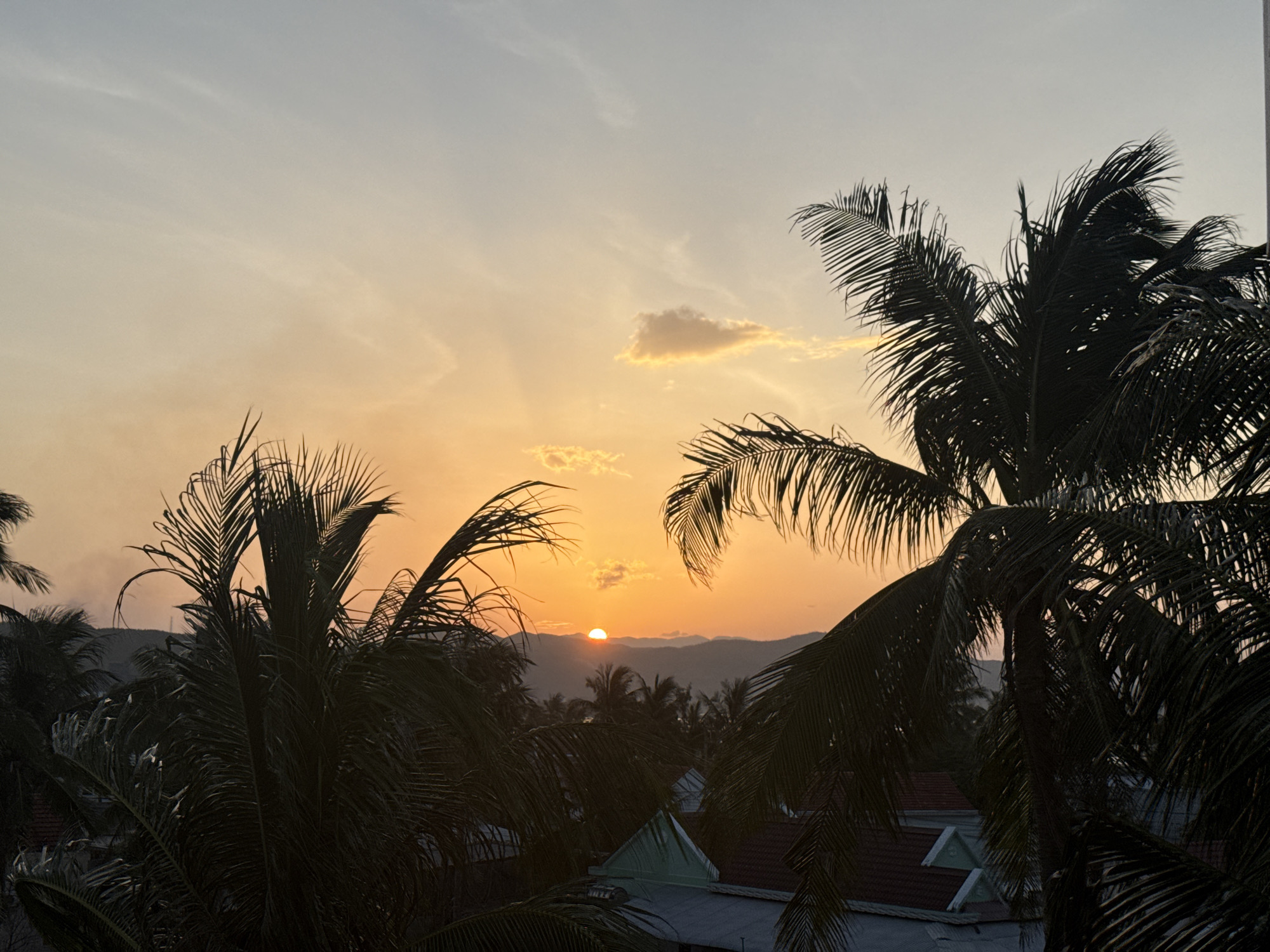 This image captures a sunset over a tropical landscape. In the foreground, the dark silhouettes of palm trees frame the scene. The sky is a gradient of colors, transitioning from a soft orange near the horizon to a pale blue at the top. A few scattered clouds are illuminated by the setting sun, which is partially visible behind a range of mountains in the distance. The mid-ground consists of more palm trees and rooftops of buildings, suggesting a settlement within this tropical environment. The overall atmosphere is tranquil and serene, characteristic of a tropical evening.