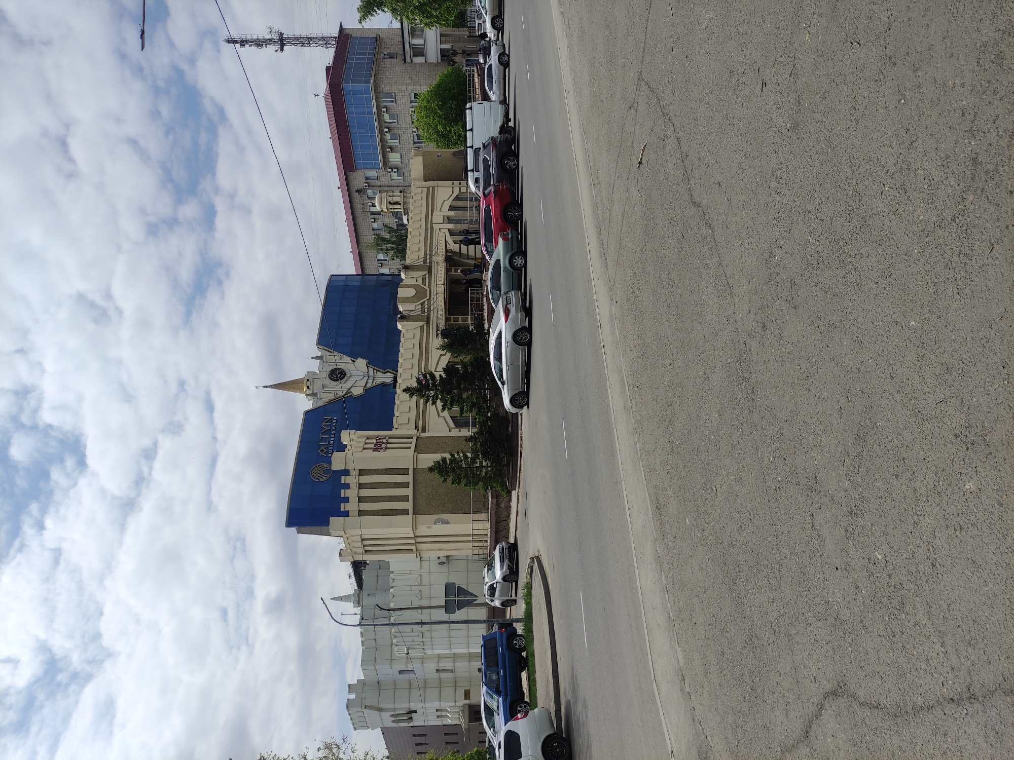 The image depicts a street scene in Pavlodar, Kazakhstan. On one side of the street stands the "Altyn Business Park" building, identifiable by its distinctive blue and gold facade. Cars are parked along the roadside in front of the building. The road itself is gray asphalt, showing some wear. The sky above is partly cloudy. Overhead wires are visible stretching across the scene. Further down the street, a taller building with a red roof and a telecommunications tower can be seen.