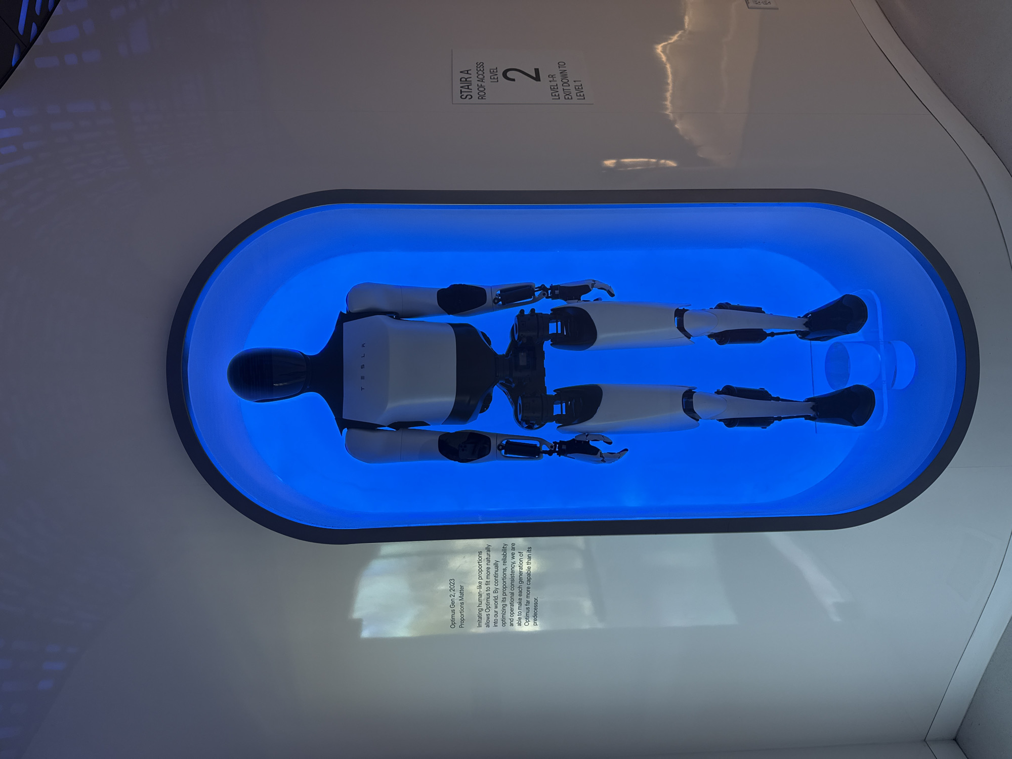 This image captures a static display of a Tesla Optimus Gen 2 humanoid robot in an indoor setting, likely a showroom or exhibition space in West Hollywood, United States of America. The robot is presented lying horizontally within a sleek, recessed, oval-shaped display case embedded in a light-colored wall. The case is dramatically illuminated from below by a vibrant blue light, giving the display a futuristic and prominent appearance.
The Optimus Gen 2 robot itself has a minimalist design, with a black head and a predominantly white body, featuring articulated limbs. The "TESLA" logo is subtly visible on its torso. The robot appears pristine and advanced.
Accompanying the display is a block of text, which reads:
"Optimus Gen 2, 2023
Proportions Matter
Imitating human-like proportions
allows Optimus to fit more naturally
into our world. By continually
optimizing its proportions, reliability
and operational consistency, we are
able to make each generation of
Optimus far more capable than its
predecessor."
Further to the right, a white sign with black text indicates directional information for the building:
"STAIR A
ROOF ACCESS
LEVEL
2
LEVEL 1-R
EXIT DOWN TO
LEVEL 1"
The wall surrounding the display is uniform and light-toned, with some reflections visible on its glossy surface, suggesting an artificial lighting environment.