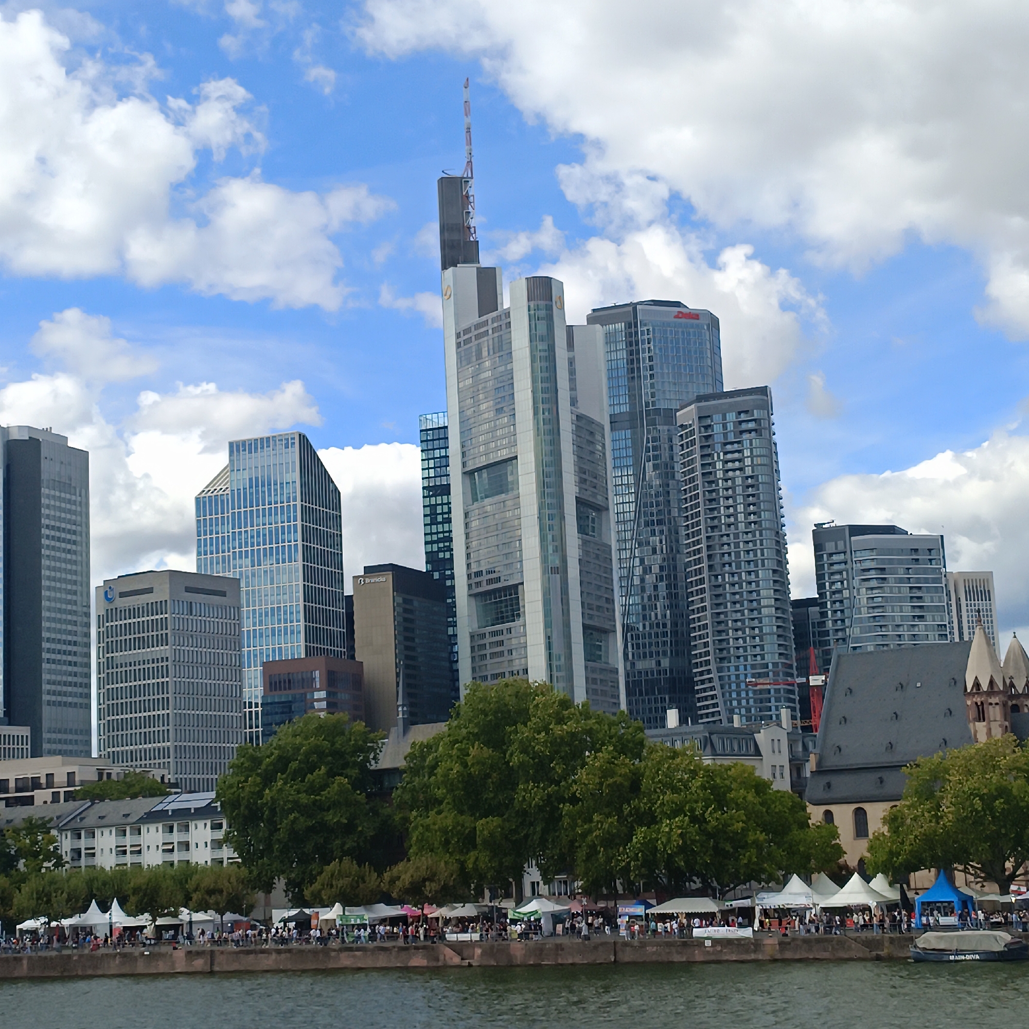 The image depicts a waterfront scene in Frankfurt, Germany, with a distinct skyline of modern skyscrapers and a historic church. In the foreground, a body of water reflects the cloudy sky and the buildings. Along the waterfront, a row of white and colored tents suggests an outdoor market or festival, with numerous people gathered, walking, and interacting. Trees line the area behind the tents, separating the waterfront activity from the imposing city architecture.
The skyline is dominated by several tall, contemporary buildings, including the Commerzbank Tower with its distinctive antenna. To the right of the modern structures, a gothic-style church with spires stands out, providing a stark contrast to the surrounding high-rises. The sky is a mix of blue and white clouds, indicating daytime. The overall impression is a bustling urban environment where modern architecture and historical elements coexist, with a lively public event taking place along the river.