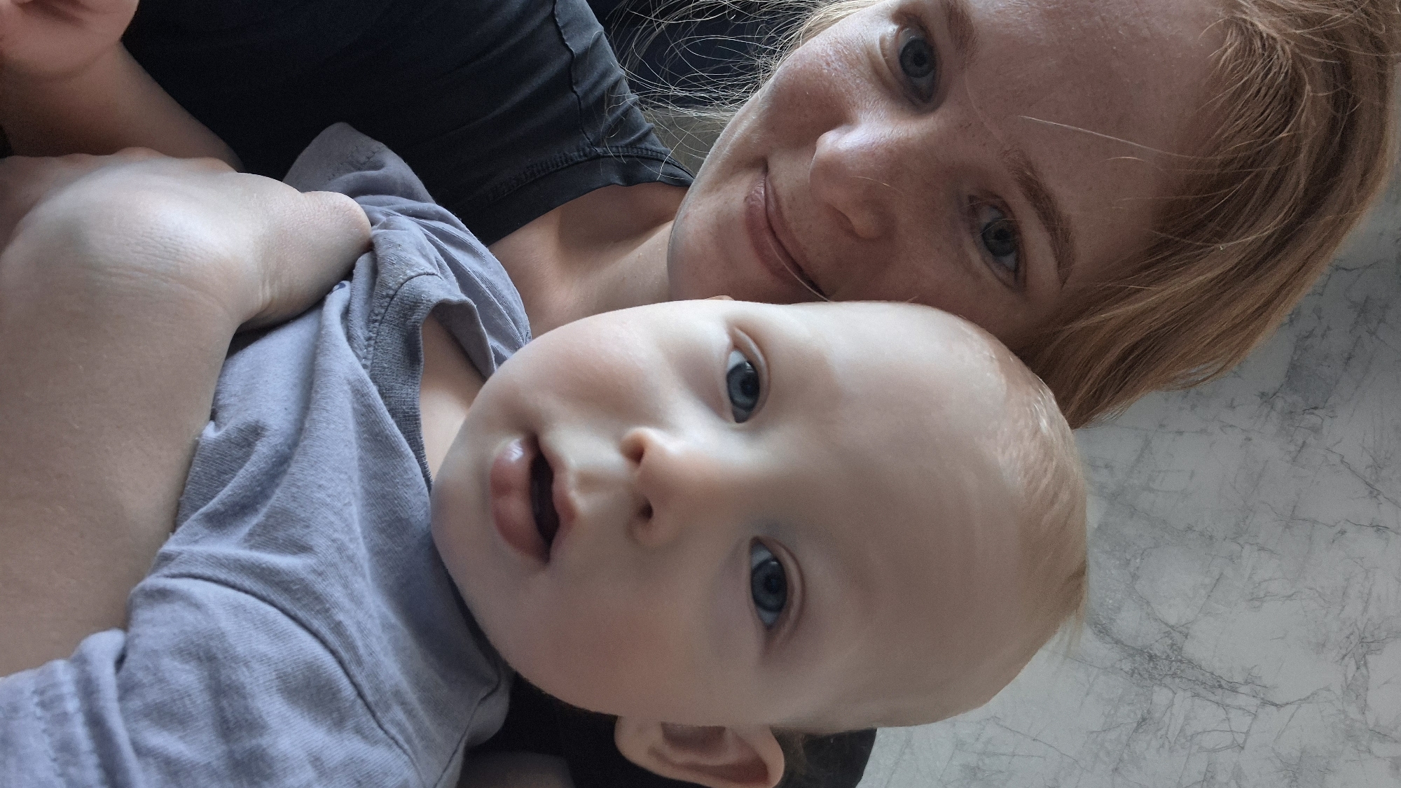 A woman with fair skin and red hair holds a baby in her arms. Both the woman and the baby have blue eyes. The baby is wearing a gray t-shirt. The woman is wearing a dark gray shirt. The background appears to be a light gray surface with a mottled pattern, possibly a marble countertop. The location is not discernable from the image.