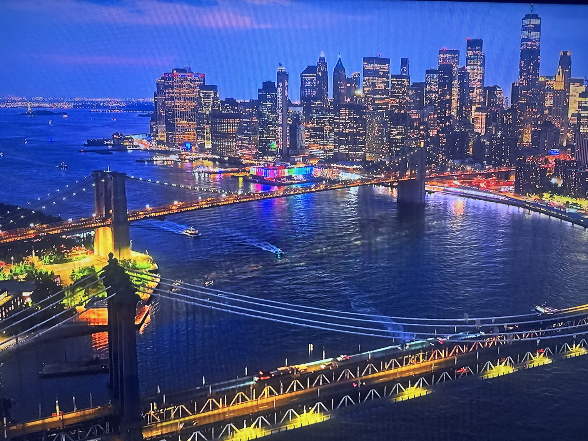 The image shows a nighttime view of the New York City skyline with the Brooklyn and Manhattan Bridges spanning the East River. The skyline is illuminated with numerous lights from buildings, and the bridges are adorned with lights highlighting their structures. Traffic is visible on the bridges. The river reflects the city lights, adding to the overall illumination. The time of day appears to be dusk or night.