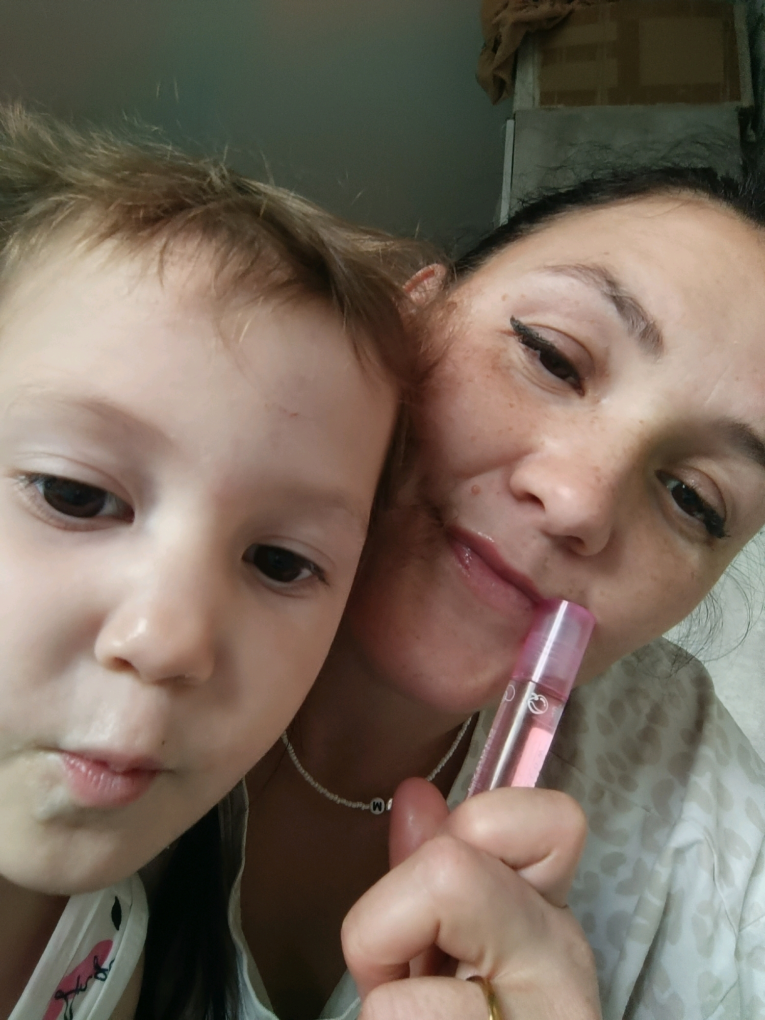 A close-up, selfie-style photograph shows a woman and a young child together. The woman, on the right, has dark hair and is looking towards the camera with a slight smile. She is holding a pink lip gloss applicator near her mouth. Her face is marked with freckles. The child, on the left, has lighter hair and is looking downwards. Both individuals are close together, suggesting an intimate moment. The background is indistinct but appears to be indoors, with a hint of shelving and draped fabric visible. No specific landmarks of Chaikovsky, Russia, are discernible in the image.