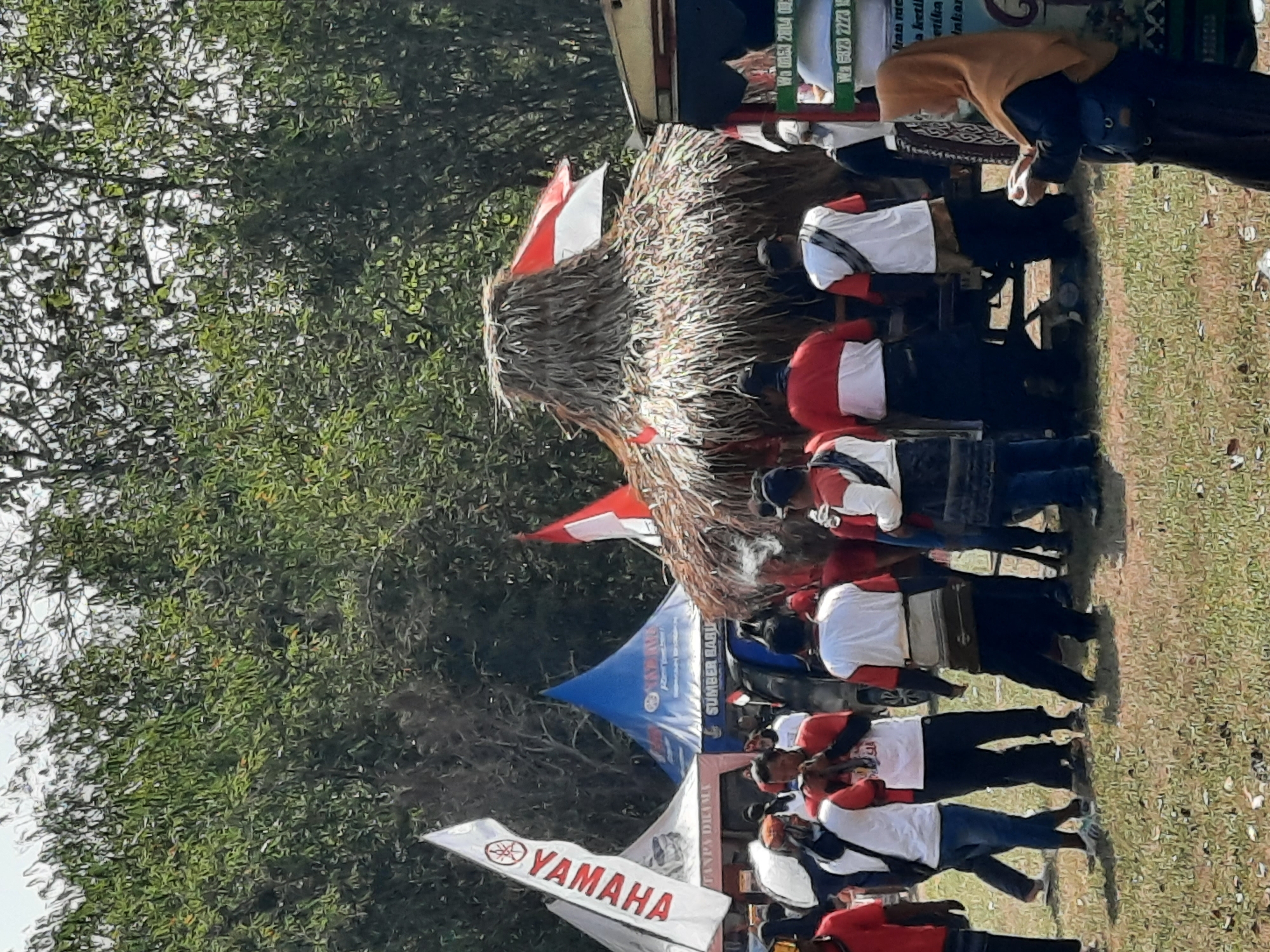 This image depicts an outdoor event with several tents, a large thatched-roof structure, and numerous people in the city of Lugu, Indonesia. The scene appears to be a community gathering or festival.
In the foreground, several individuals are standing and interacting. Some are wearing white shirts with red accents and dark pants, while others are in casual attire. A woman in a brown hijab and dark dress is visible on the right, looking down at something in her hands. Behind them, a large, conical thatched-roof structure, resembling a hut or temporary stage, is the central focus. Two Indonesian flags (red and white) are attached to this structure, and another red and white flag is seen on a pole to the right.
In the mid-ground, various tents are set up. One white tent prominently displays a large "YAMAHA" logo with a red tuning fork emblem. Another tent, blue and cone-shaped, also has a "YAMAHA" logo and text that reads "SUMBER BARU." Other tents are visible further back. A dark-colored vehicle is partially visible behind the thatched-roof structure.
The environment is an open, grassy field, with dense green trees forming a backdrop. The sky is overcast or hazy. The overall impression is of a lively outdoor event or market.