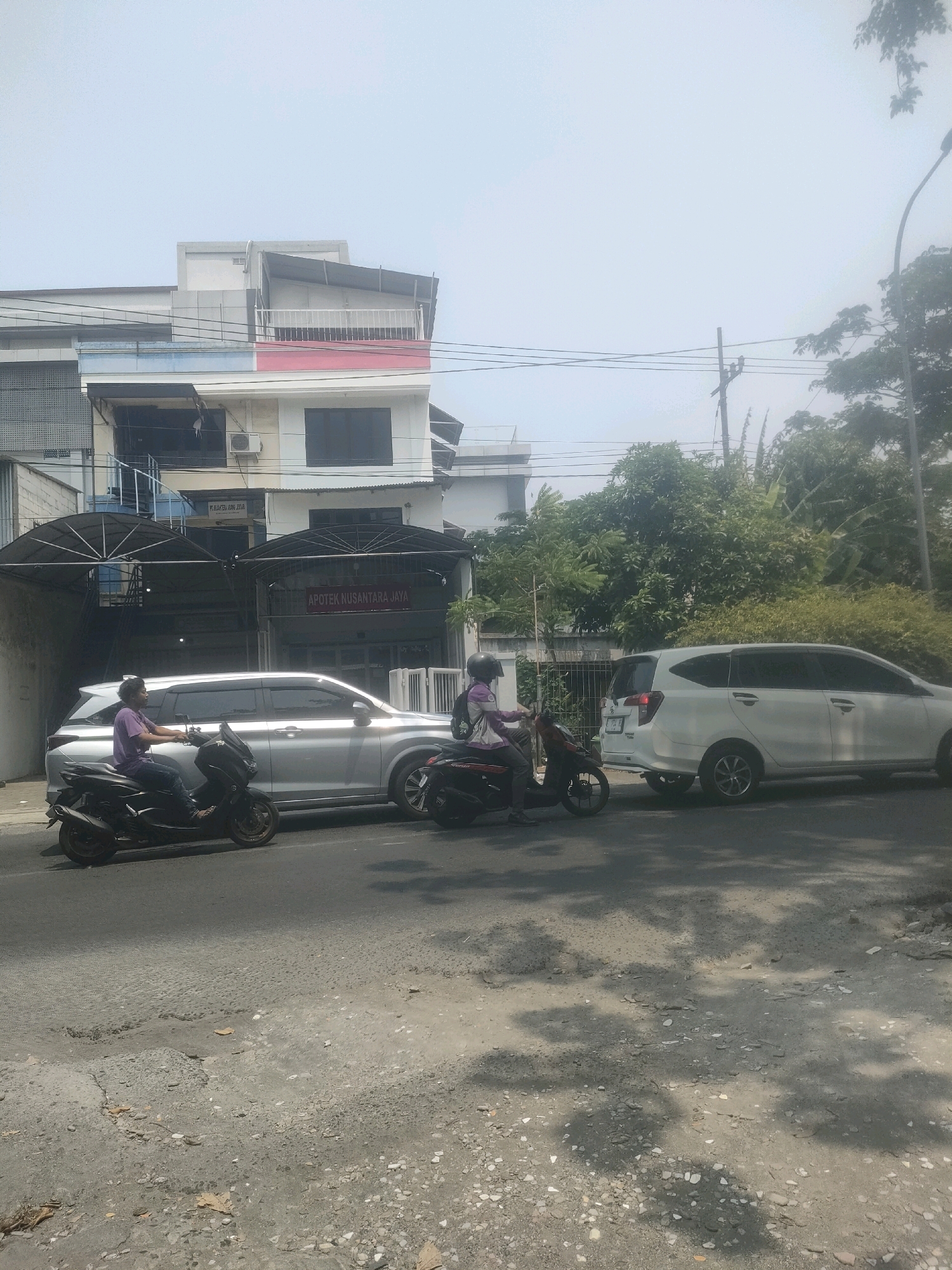The image depicts a street scene in Surabaya, Indonesia, on a bright, hazy day. In the foreground, a damaged asphalt road with visible potholes and scattered dry leaves indicates a state of disrepair. Two motorcycles are present on the road. The motorcycle on the left is black, ridden by a person wearing a purple t-shirt and dark pants, with their back to the camera. The motorcycle on the right is also black and is ridden by a person wearing a helmet, a jacket, and pants, also with their back to the camera, their left arm resting on the handlebars.
Alongside the road, a silver SUV is parked. Further down the street, a white minivan is visible, also on the road. Behind the vehicles, a multi-story building dominates the background. The building features a section with a red and white facade, indicative of the Indonesian flag's colors. Above this, there are more stories with windows and a balcony. A prominent sign on the building reads "APOTEK NUSANTARA JAYA," identifying it as a pharmacy. Electrical wires are strung across the scene, and trees with dense foliage are present on the right side of the image. The sky is overcast and bright, suggesting a sunny but humid atmosphere typical of Surabaya.