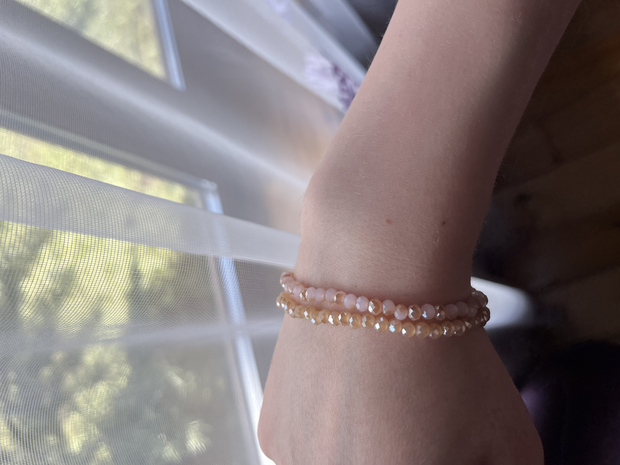 The image shows a close-up of a person's forearm and wrist, adorned with two stacked, delicate bracelets made of small, faceted beads in shades of light pink and pale gold. The bracelets are positioned around the wrist. The background is softly blurred, revealing a sheer white curtain with a hint of green foliage visible through it, suggesting an indoor setting near a window. The lighting appears to be natural daylight, casting a soft glow on the skin and bracelets. There is no information in the image to indicate the location in the city of Ashkadar, Russia.