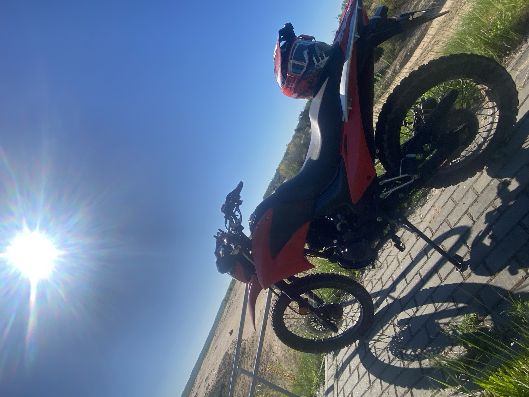 The image shows a red and black dirt bike parked on a brick surface with a helmet on the back. The location is Парэцкі сельскі Савет, Belarus, as indicated by the presence of what appears to be a sand dune or quarry in the background. The sky is bright blue, and the sun is visible in the upper left corner of the frame, causing a lens flare. The motorcycle is angled in the image, and the kickstand is deployed to keep it upright. Grass grows along the edge of the brick surface. A metal fence is present in the background.