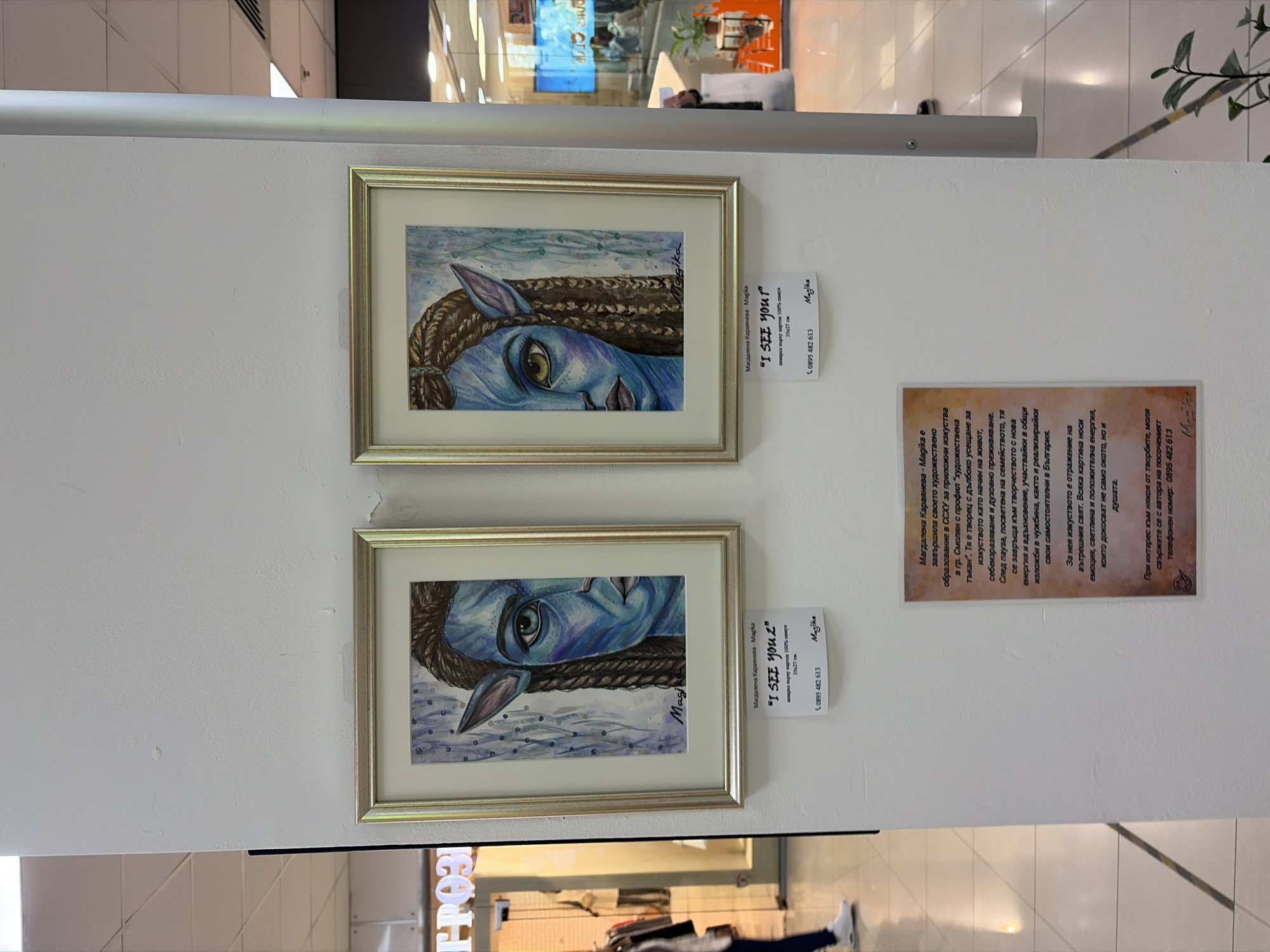 The image shows two framed watercolor paintings of a blue Na'vi face, likely from the movie Avatar. The paintings are displayed side-by-side on a white wall, and each has a tag next to it with the title "I SEE YOU?" and the artist's name, Магдалена Караянева - Magika, along with contact information. There's a larger text block beside the paintings with a description of the artist. The setting appears to be an indoor space, possibly a shopping mall or gallery, as suggested by the presence of other stores visible in the background.