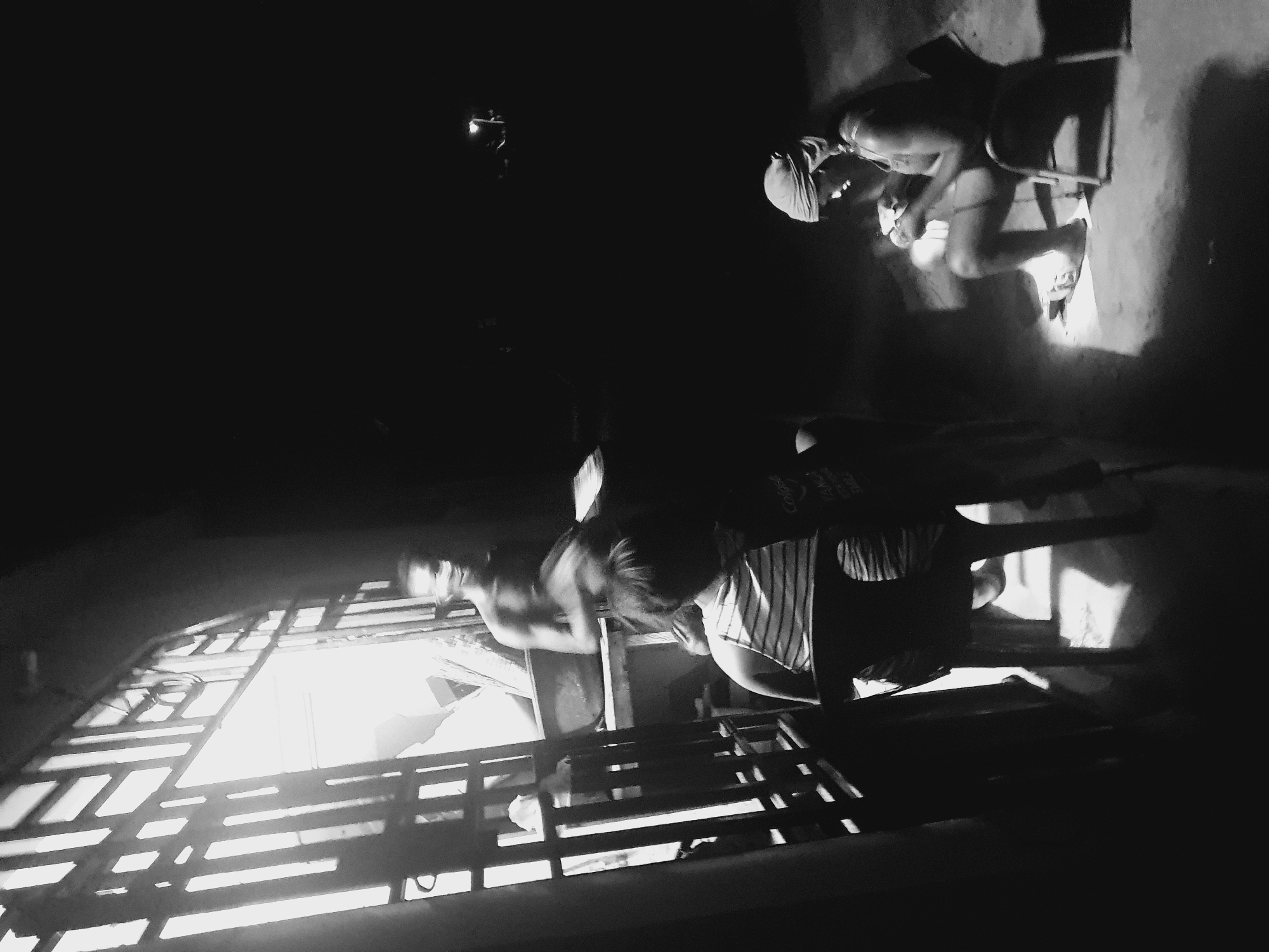 A low-angle, black and white photograph depicts a partially constructed structure in what appears to be an interior space. Light streams through the unfinished wooden framework, illuminating areas within. Two individuals are visible: one is seated on a chair and possibly working with an object, while another stands nearby. The location is Lokoja, Nigeria.