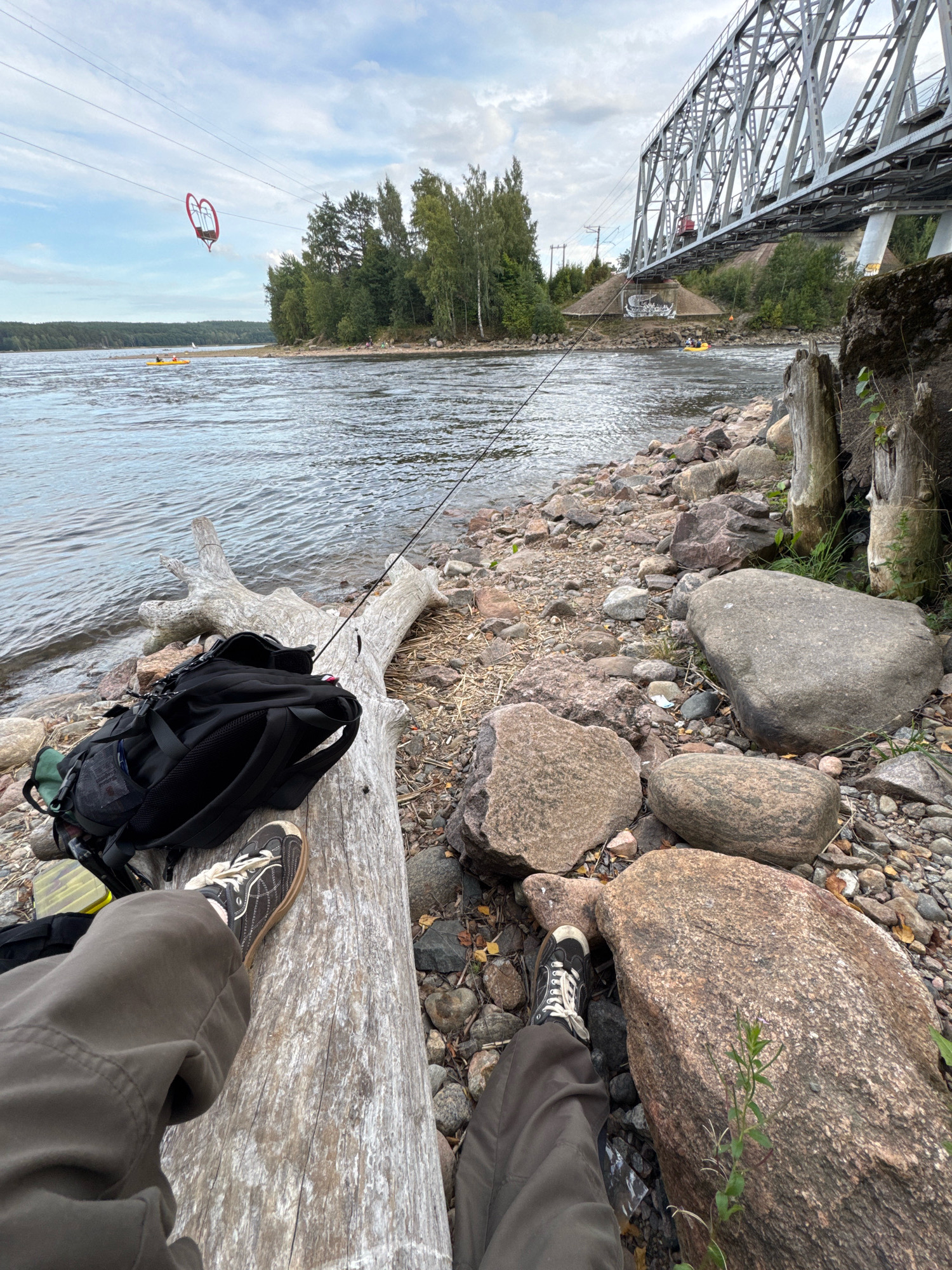 The image depicts a scene on the rocky shore of a river, with a large railway bridge spanning the water in the background. In the foreground, a person's legs and feet, clad in dark pants and canvas sneakers, are resting on a weathered, light-colored log. A black backpack is placed on the log next to the person's legs, and a fishing rod extends from the log towards the water. The river is calm, with a gentle ripple on its surface. In the middle distance, two yellow kayaks can be seen on the water, with a heart-shaped structure suspended from overhead wires further in the background. The environment is natural, with trees lining the opposite bank of the river and large rocks scattered along the shore. The sky is cloudy, suggesting a temperate climate. The location is identified as Ovragi, Russia.