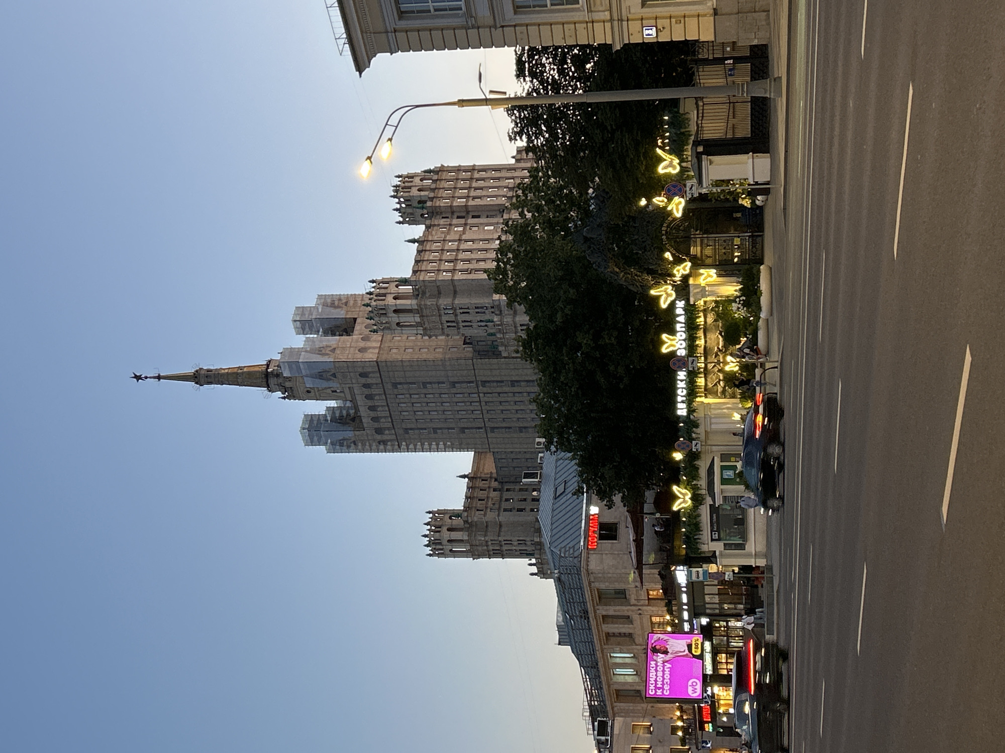 This is a low-angle, medium shot of the Moscow State Circus Building (also known as the Nikulin Circus) and surrounding buildings, taken at dusk. The sky is a clear, pale blue. In the background, the large, ornate circus building dominates the frame with its distinctive spires and architectural details, appearing to be under some form of renovation or scaffolding on its upper sections. To the left of the circus building, a lower, more modern-looking building houses shops and restaurants, with illuminated signage including a bright pink advertisement. In the mid-ground, a tree-lined area features an entrance gate decorated with illuminated butterfly-shaped lights, and signage that reads "ДЕТСКИ ЗООПАРК" (Children's Zoo Park). A lamppost with two lights stands in the mid-right, casting a warm glow. In the foreground, a multi-lane asphalt road shows white lane markings, and two dark-colored cars are in motion, with their taillights illuminated. Several people are visible on the sidewalk and crossing the street in the mid-ground. The overall scene depicts an urban environment transitioning from day to night, with a mix of historic and contemporary architecture.