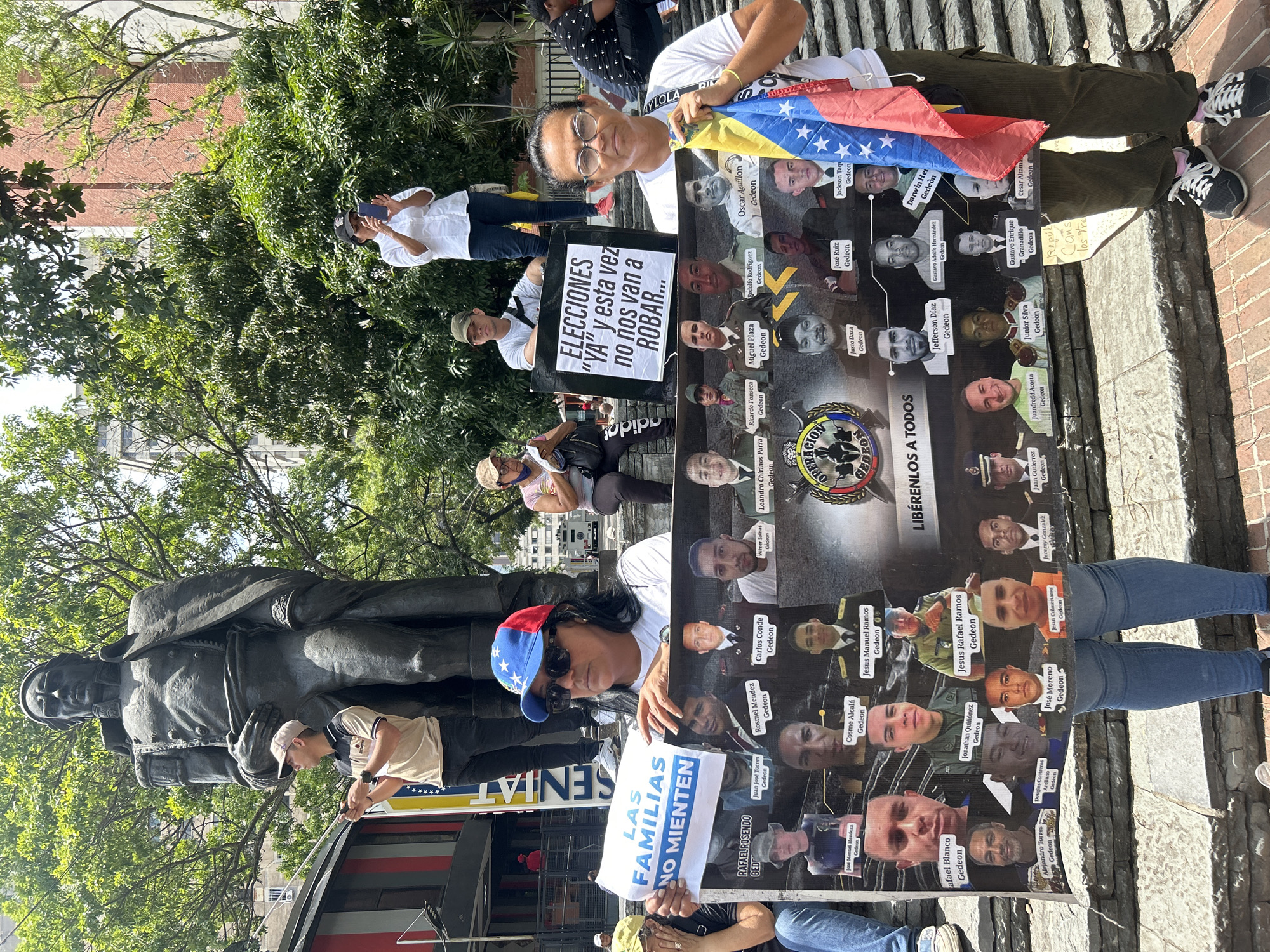 The image depicts a public protest taking place in an urban square in Caracas, Venezuela, on a sunny day. Several individuals are gathered, holding signs and a Venezuelan flag, indicating a demonstration of political or social advocacy.
In the foreground, a woman wearing glasses and a white shirt holds a large, prominent poster featuring multiple headshots of men, many identified with "Gedeon" next to their names. The poster also displays a logo that reads "OPERACIÓN GEDEON" and a bold message at the bottom, "LIBÉRENLOS A TODOS" (Free them all), suggesting a demand for the release of these individuals, likely related to the 2020 "Operation Gedeon" incident. She also holds a Venezuelan flag. Beside her, another person in a cap holds a smaller, rectangular sign that reads: "ELECCIONES 'YA' y esta vez no nos van a ROBAR..." (Elections "NOW" and this time they won't ROB us...), pointing to a demand for immediate elections and an accusation of past electoral fraud or corruption. To the left, a person wearing a blue, red, and white cap holds a sign stating: "LAS FAMILIAS NO MIENTEN" (Families don't lie).
In the background, a large, dark statue of a man, likely a historical figure, stands among green trees, characteristic of a public park or plaza. Other people are visible in the background, some observing or taking photos with their phones. The overall scene suggests a determined, peaceful protest calling for political change and justice. Visible text includes "SENIAT" partially seen on a structure and "MY LOLA RIV" on a shirt in the background.