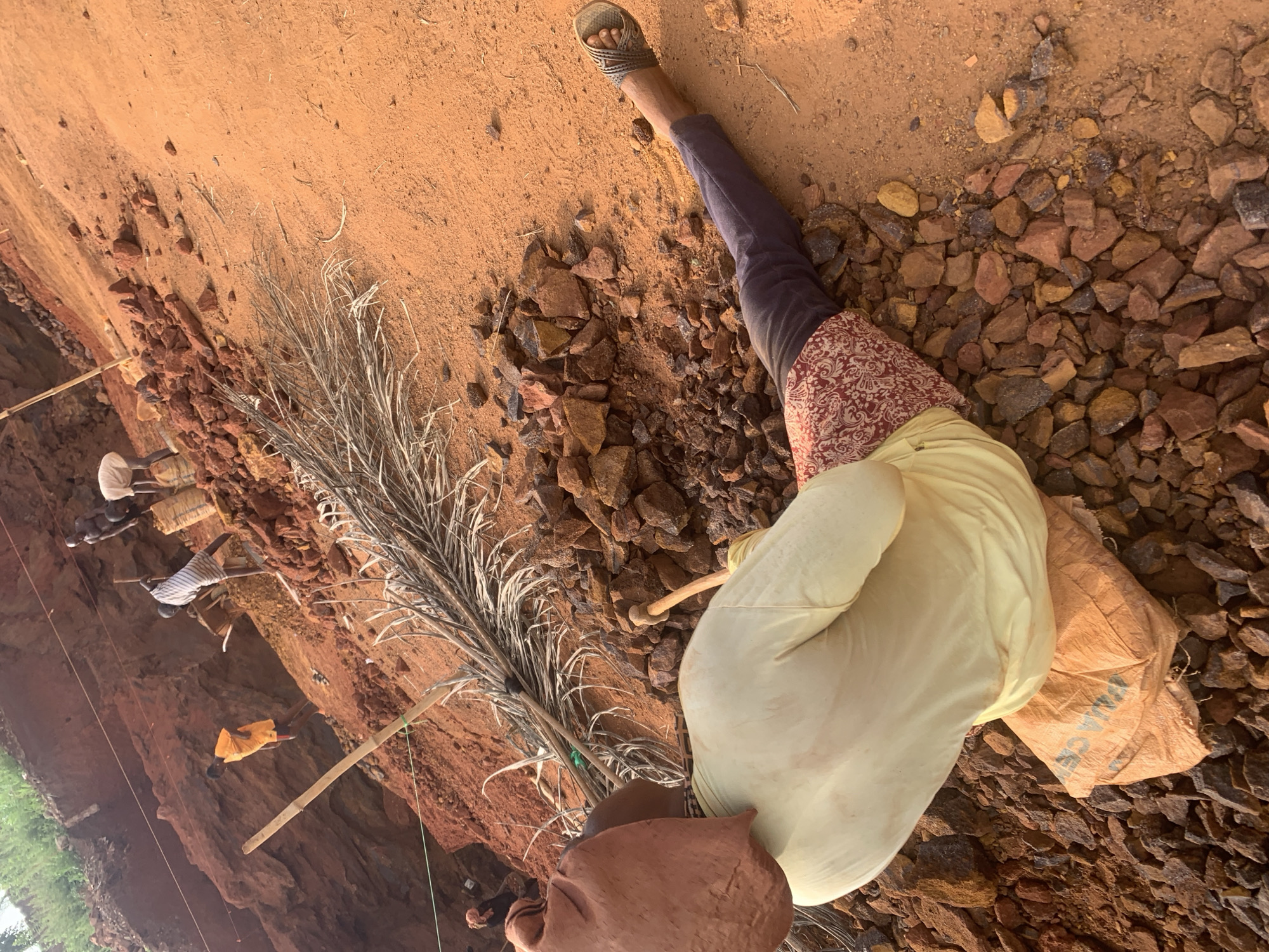 An image depicts a scene in Ukwu-Abwa, Nigeria. A person wearing a light yellow top, a patterned wrap, and dark pants with a sandal on their foot is standing on rocky, reddish-brown terrain. They are holding a bundle of dry, palm-like leaves tied together with a rope. A few other figures are visible in the distance near what appears to be a dirt wall or cliff. Bags of something sit on the rocks near the person, and various stakes and ropes are present in the scene.