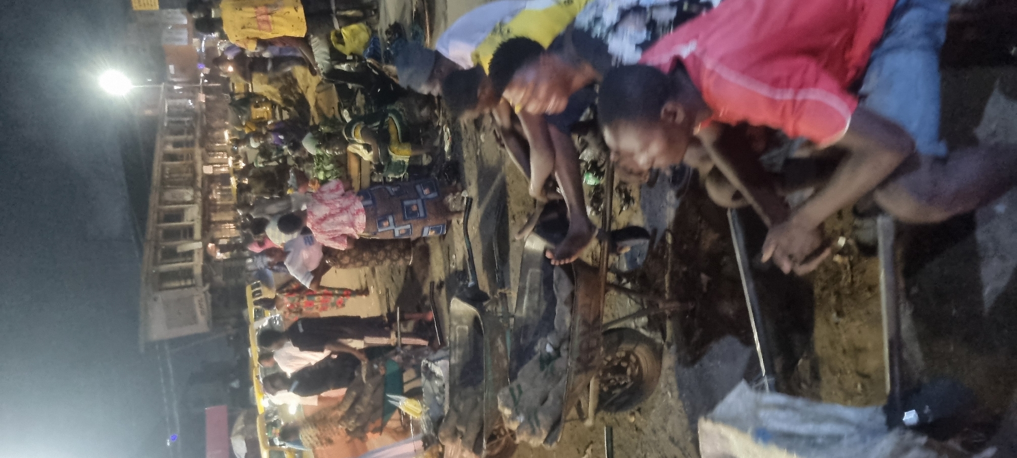 The image captures a scene in Lagos, Nigeria, focusing on a group of people gathered around what appears to be a damaged or disabled vehicle. The location seems to be an open area with streetlights in the background, suggesting an urban setting, possibly a roadside or a marketplace.
A crowd of people are visible in the background, and several individuals are closely inspecting and working on the vehicle. The lighting conditions suggest it is either late evening or night. The foreground highlights the individuals directly involved with the vehicle. Their actions seem to be focused on assessing the damage and potentially attempting repairs.