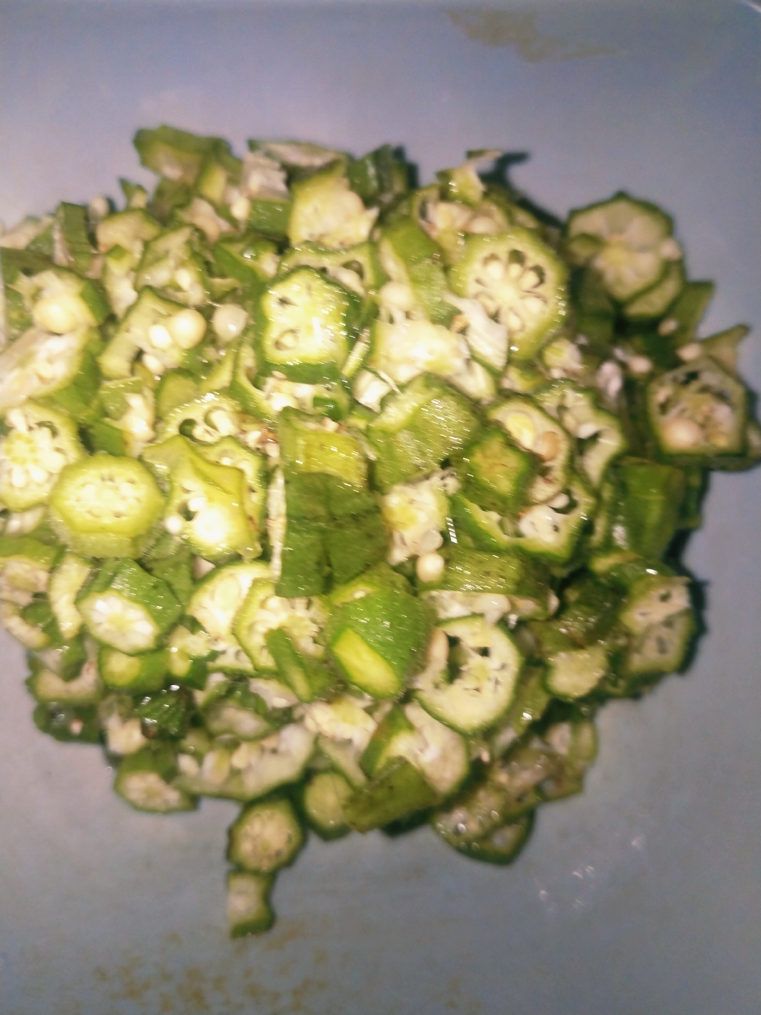 A pile of chopped okra fills a light blue bowl, which is set against a blurred light blue background. The okra pieces are cut into small, round slices with visible seeds in the center. The pieces are a vibrant green and appear slightly wet, suggesting they may have been washed or are about to be cooked. The lighting is dim, and the image is slightly out of focus, giving it a soft, natural appearance. There are no people or animals visible in the image. The scene appears to be indoors, likely in a kitchen.