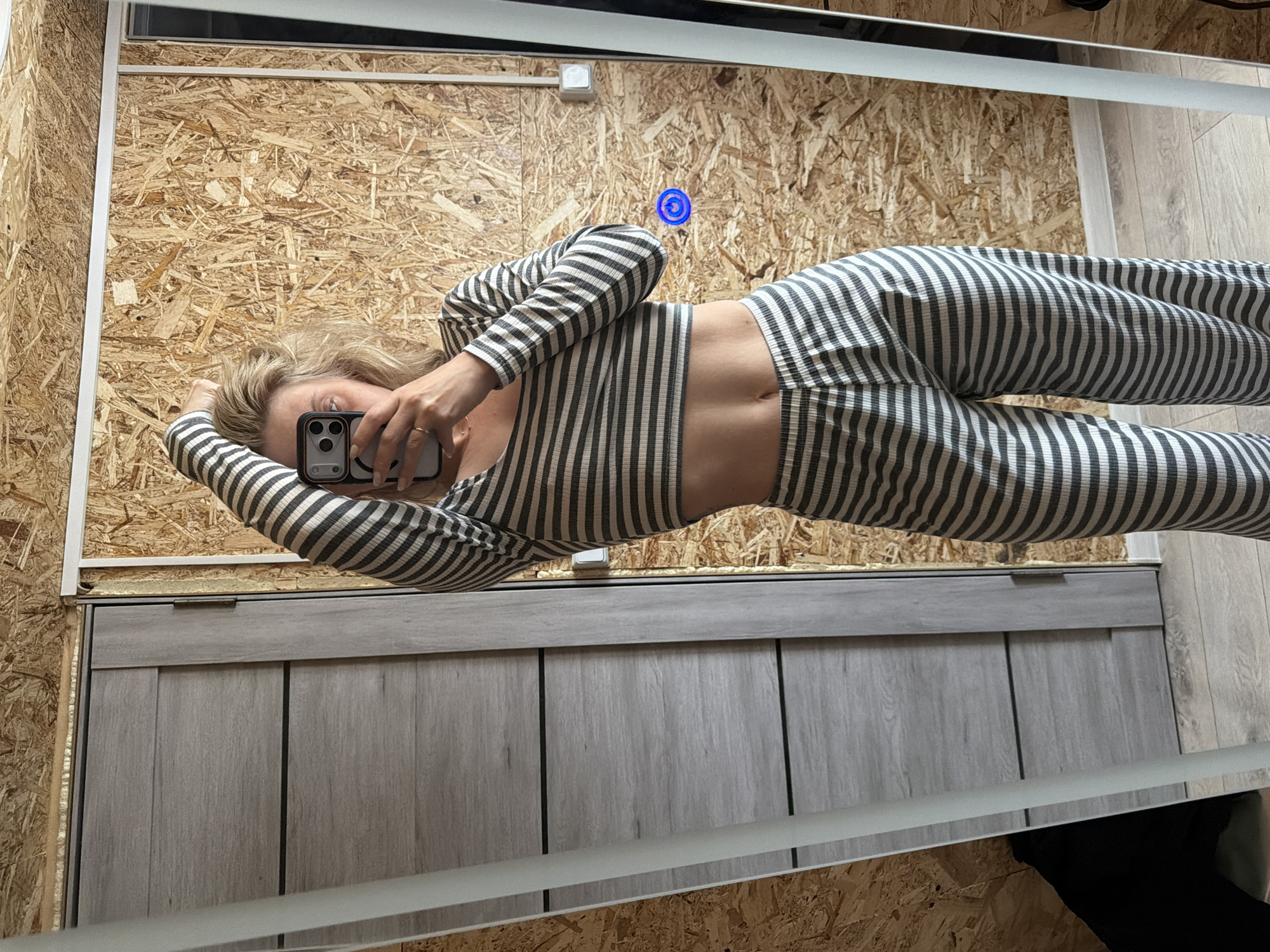 A woman is taking a mirror selfie. She is wearing a striped long-sleeved shirt and pants, and is holding a smartphone in her right hand. The background consists of a wall constructed of oriented strand board (OSB) and a bathroom cabinet. The reflections in the mirror suggest the scene may be located in a bathroom. There is no evidence to support that this image was taken in the city of Chaikovsky, Russia.