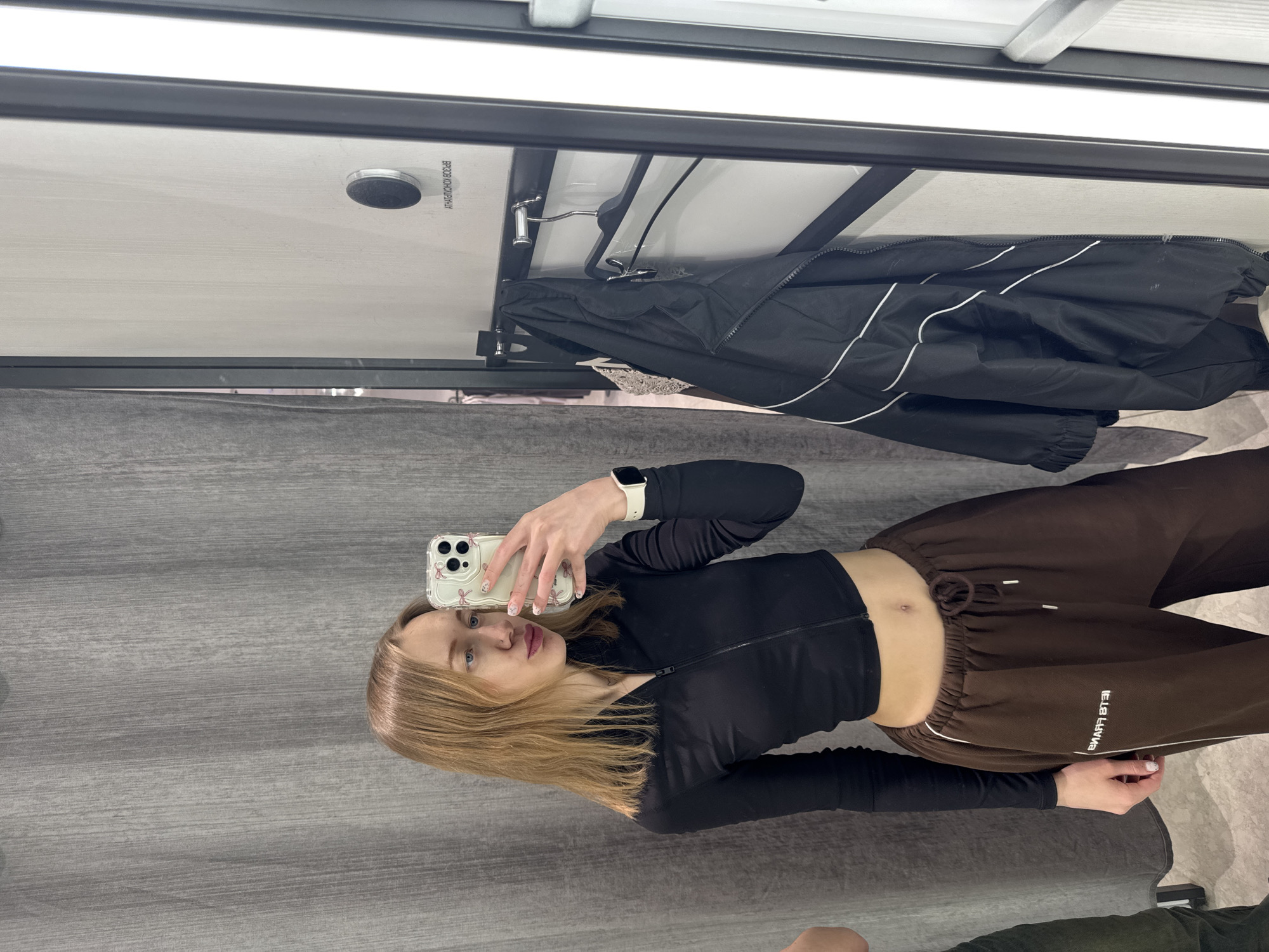 A young woman is taking a selfie in what appears to be a fitting room, with a gray curtain in the background. She is wearing a black long-sleeved crop top and brown sweatpants. She is holding a phone with a clear case, visible in the mirror's reflection. A black jacket hangs on a rack in the background, along with part of the fitting room's structure. The location is not Rostov-on-Don, Russia, and there is no information that places the image there.