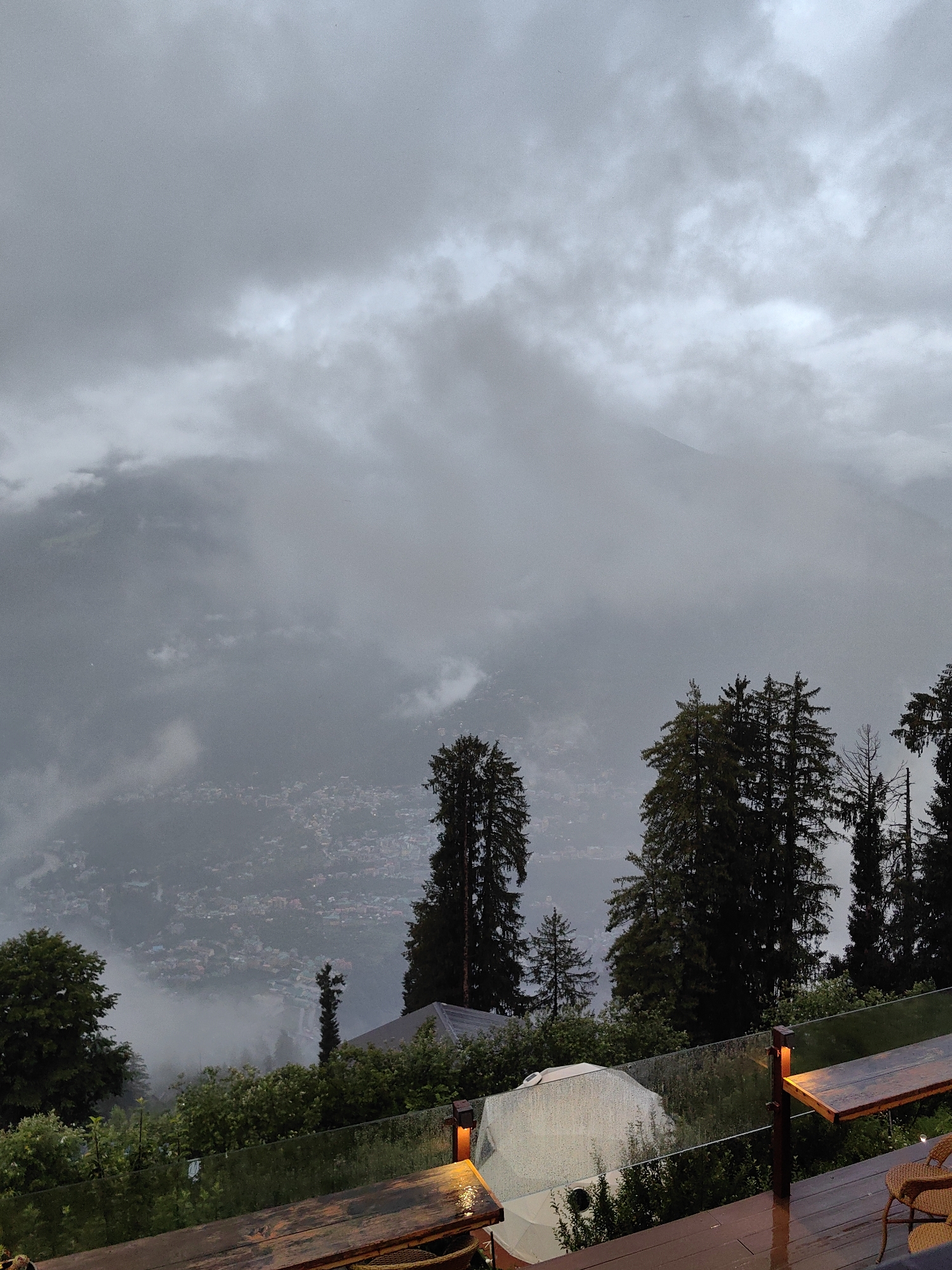 The image depicts a misty, overcast view of the town of Prini, India, from a slightly elevated vantage point. In the foreground, a wooden deck with a glass railing is visible, with a wooden table and a woven chair occupying the right side of the frame. The deck surface appears wet, suggesting recent rain.
The middle ground is characterized by lush green foliage and several tall pine trees. A white tent-like structure is partially obscured by the greenery. Beyond this, the town of Prini unfolds, its buildings clustered together and appearing somewhat hazy due to the mist. The distant landscape is dominated by mountains shrouded in thick clouds, creating a dramatic and atmospheric scene. The overall impression is one of a tranquil, albeit slightly somber, mountain setting during a cloudy day. There are no people visible in the image.