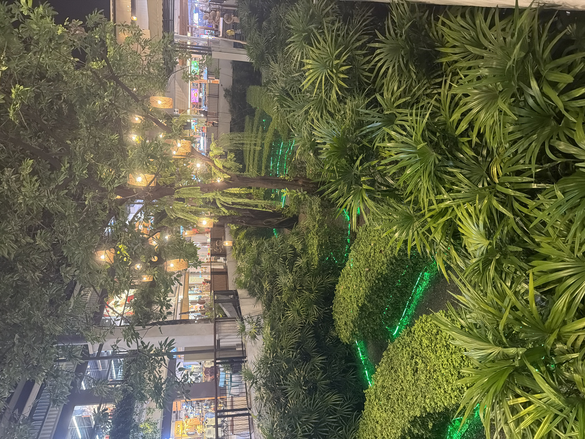The image depicts a nighttime view in what appears to be an outdoor area within a multi-level commercial or retail complex. This area is located in บ้านบางรักษ์, Thailand. The foreground is dominated by dense, green vegetation, including various types of plants and bushes. Some of the bushes have bright green LED lights outlining their shapes. A tree with illuminated lamps hanging from its branches is visible. In the background, multiple levels of shops and businesses can be seen, illuminated with bright lights, and people can be seen on multiple levels. The architectural style appears modern, with metal railings and glass windows. The overall lighting is a mix of artificial lights from the shops and the decorative lights in the vegetation. The perspective is slightly angled, giving a view down into the garden area.