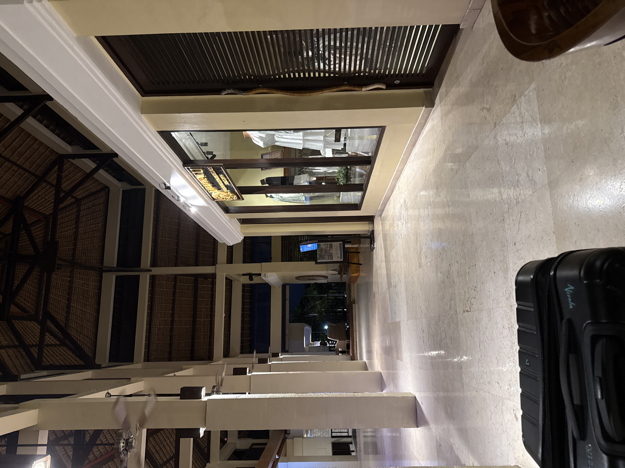 The image depicts a hotel lobby with a high, thatched roof supported by large, square pillars. The floor is made of polished marble tiles, reflecting the ambient light. In the foreground, a black suitcase with the word "Nissha" visible on its side is positioned on the floor. To the right of the suitcase, a shop window displays mannequins dressed in white and black clothing, with a gold-colored sign above that reads "BALI MANDIRA BEACH". The shop's interior is lit and shows racks of garments. Adjacent to the shop is a dark-colored door with horizontal slats. In the background, the lobby extends with more pillars and seating areas with rattan furniture visible further down. The overall atmosphere is dim, suggesting it is either early morning or evening. The location is identified as the lobby of Bali Mandira Beach Resort & Spa in Denpasar, Indonesia.