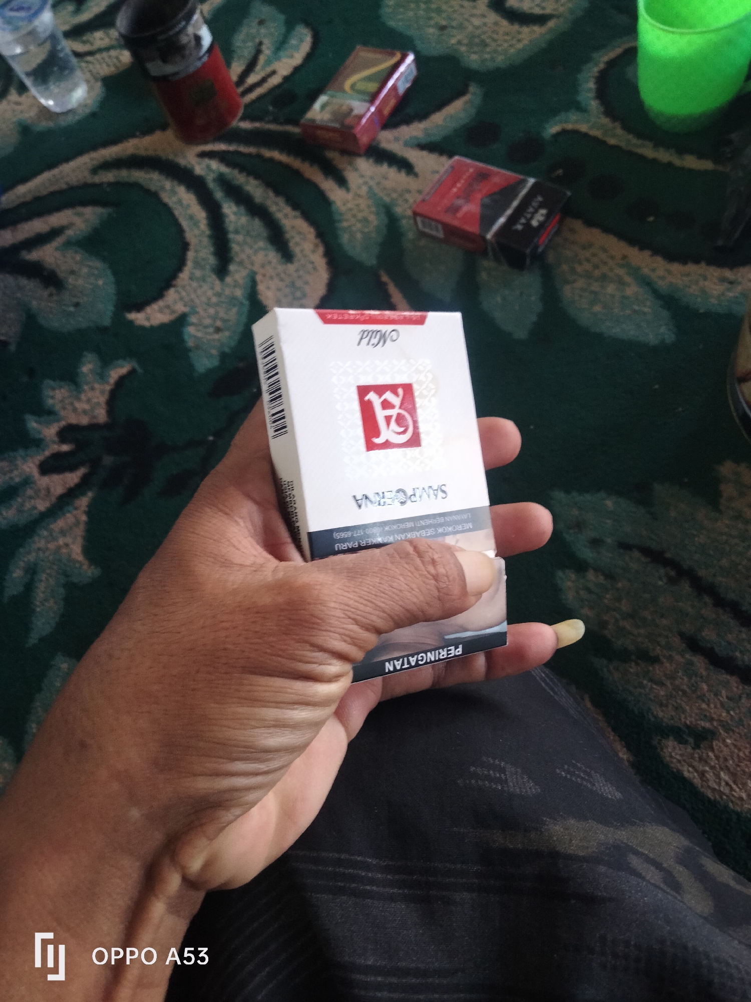 A person's hand holds a pack of Sampoerna Mild cigarettes. The pack is white with red and gold accents. The person's hand is visible from the wrist to the fingertips, with a long fingernail on the index finger. The background appears to be a patterned carpet in shades of green and brown. Other objects visible in the background include a lighter, a bottle of liquid, and another pack of cigarettes. The bottom left corner of the image has a watermark that reads "OPPO A53". The scene is not discernable to be in Badung, Indonesia.